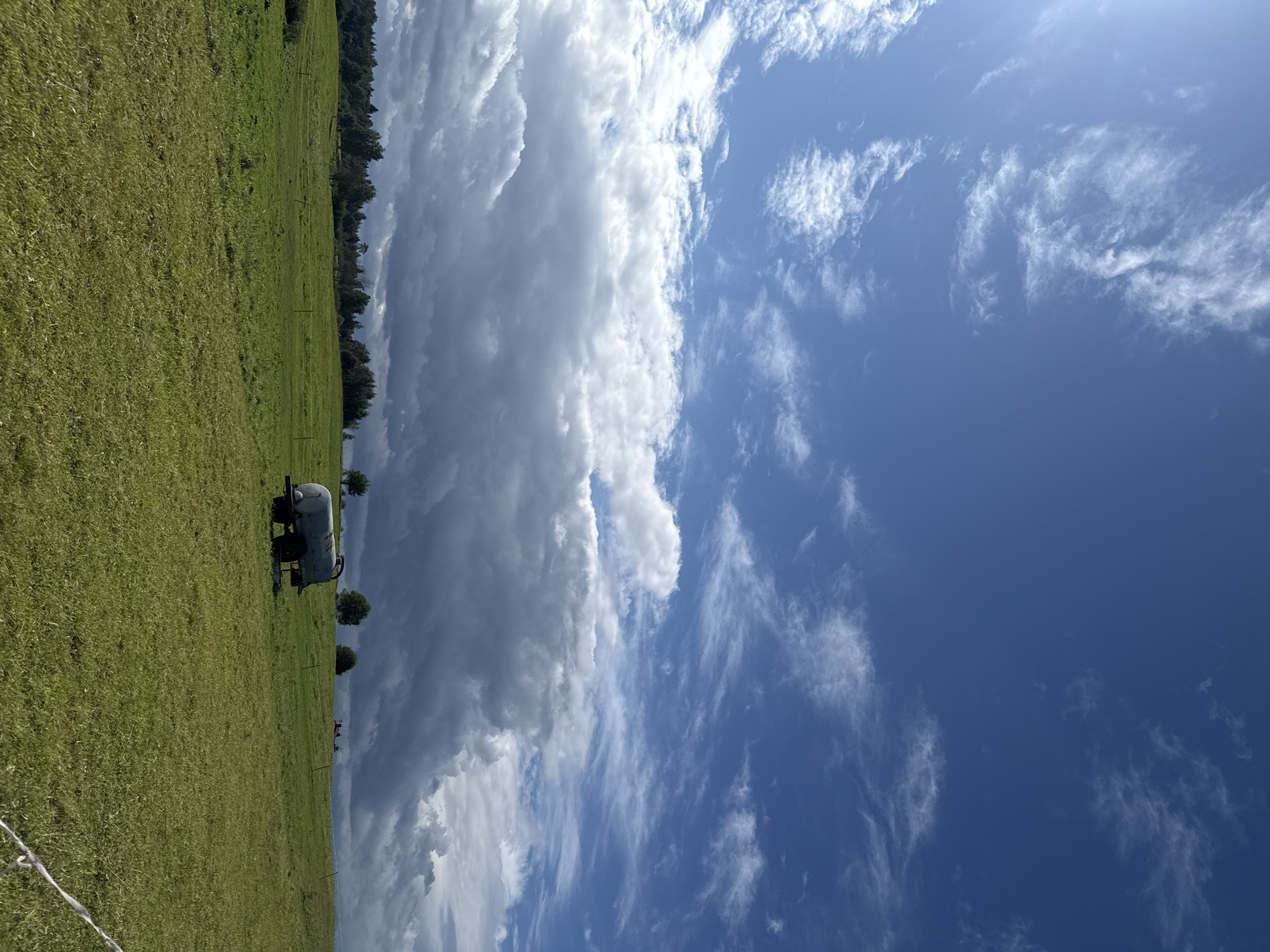 The image shows a green, grassy field in Putiniškės, Lithuania. The sky is a vibrant blue with scattered white clouds, some of which are darker and appear heavy, possibly indicating rain. In the middle ground of the field, a water tank trailer is visible, with two wheels and a cylindrical tank. Several trees are scattered across the landscape, and in the far background, a treeline suggests a wooded area. The overall impression is one of a rural, open landscape under a dynamic sky.