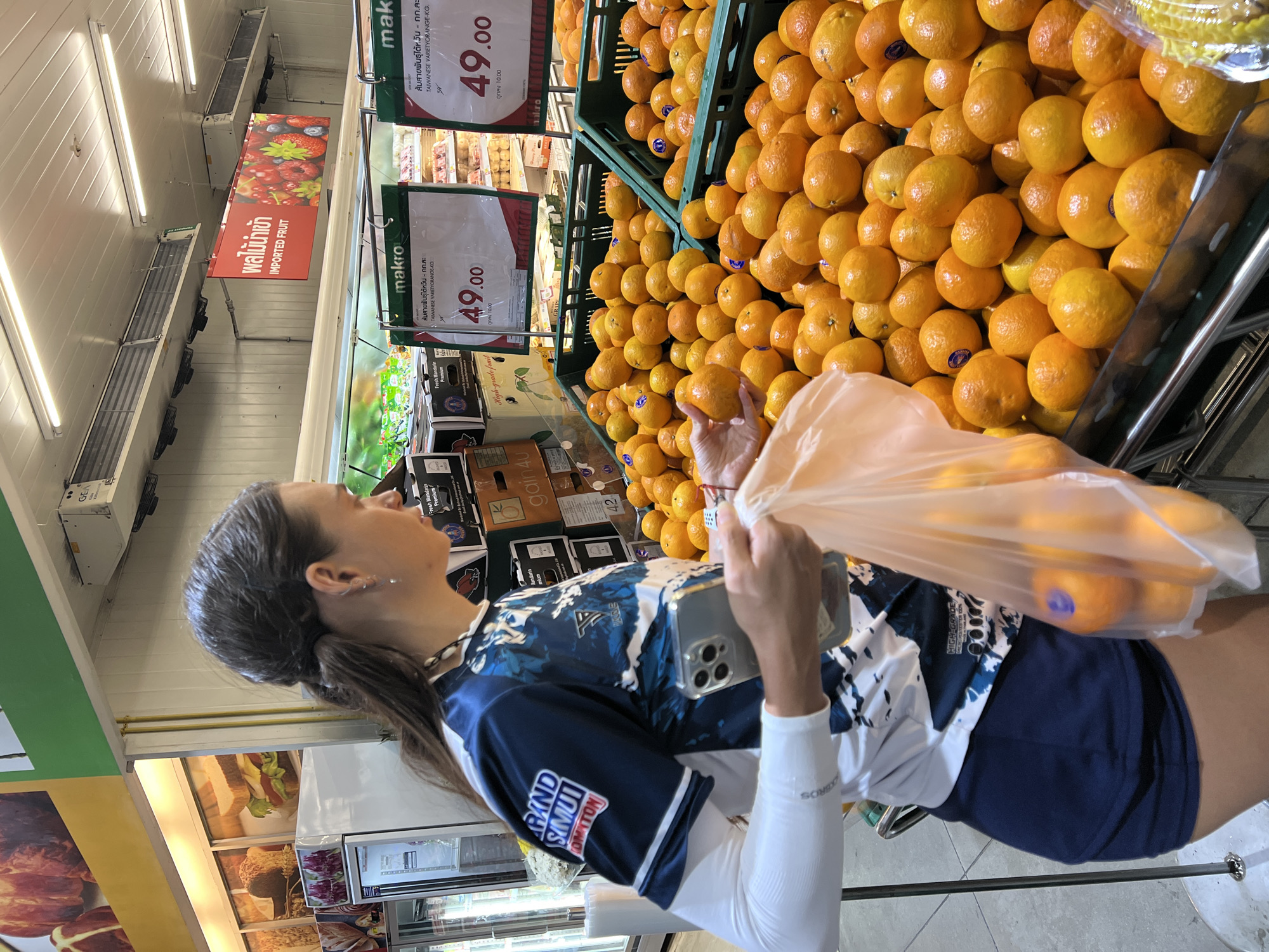 In Baan Bo Phut, Thailand, a woman is seen in a supermarket's produce section, examining a pile of fresh oranges. She is wearing a blue and white athletic top with "GRAND SIMUT" visible on the sleeve, dark blue shorts, and a white long-sleeved undershirt. Her hair is pulled back in a ponytail, and she is holding a smartphone in her right hand while reaching for an orange with her left. A translucent plastic bag, containing several oranges, is held in her left hand. The oranges are displayed in large green crates, with price tags indicating "49.00" and "TAIWANESE VARIETY ORANGE-KG." Other produce and supermarket aisles are visible in the background, including a red sign with Thai script and the words "IMPORTED FRUIT." The lighting suggests an indoor, commercial environment.