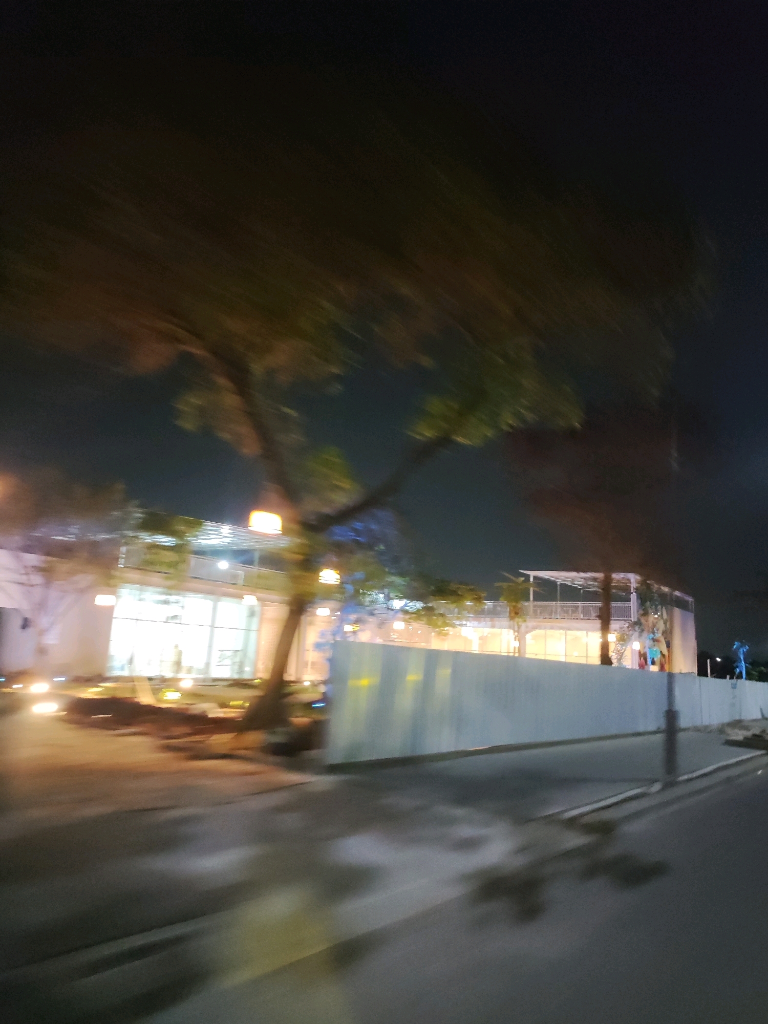 An outdoor, night-time, low-angle, full shot shows a blurred scene from a moving vehicle. The foreground is dominated by a dark, wet asphalt road with blurred reflections of lights. A light-colored, low-rise building with a large glass facade is visible in the middle ground, with lights emanating from within. A tree with blurred foliage obscures part of the building and the sky. A light-colored, corrugated metal fence runs along the front of the building, separating it from the road. A lamppost stands near the fence. In the background, the sky is dark and appears to have some faint lights. A blue statue of a person is visible on top of the fence to the right of the frame. The image is characterized by motion blur, suggesting the photograph was taken while in transit.