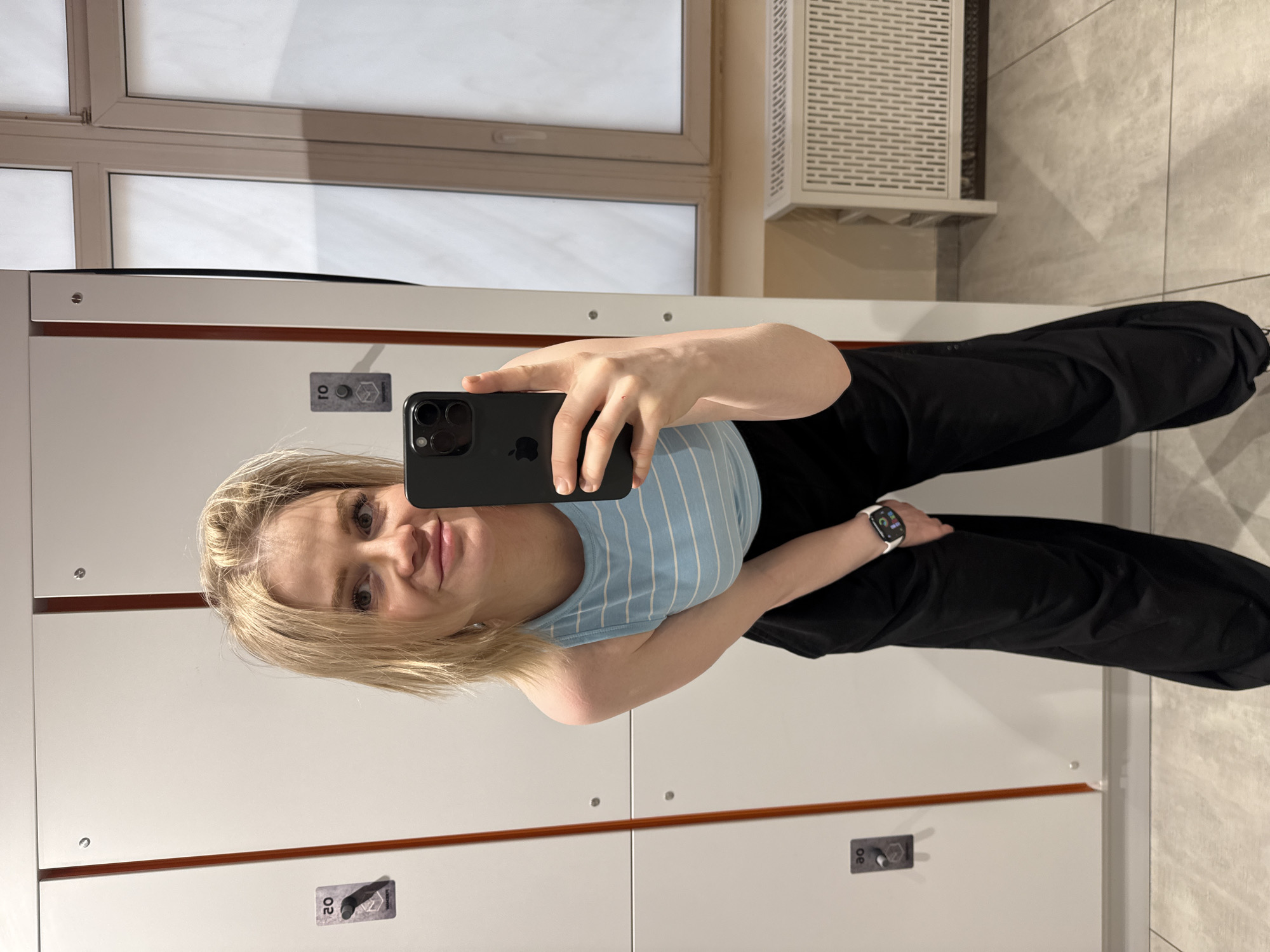 A woman is taking a selfie in what appears to be a locker room. She is wearing a blue and white striped tank top and black pants. Her blonde hair is shoulder length. She is holding a black iPhone in her hand. On her left wrist is a white smartwatch. Behind her are white lockers with silver hardware. Above the lockers, there is a window and an air conditioning unit. The background includes beige tiled walls and floor. The location is likely a gym or recreational facility in Saint Petersburg, Russia.
