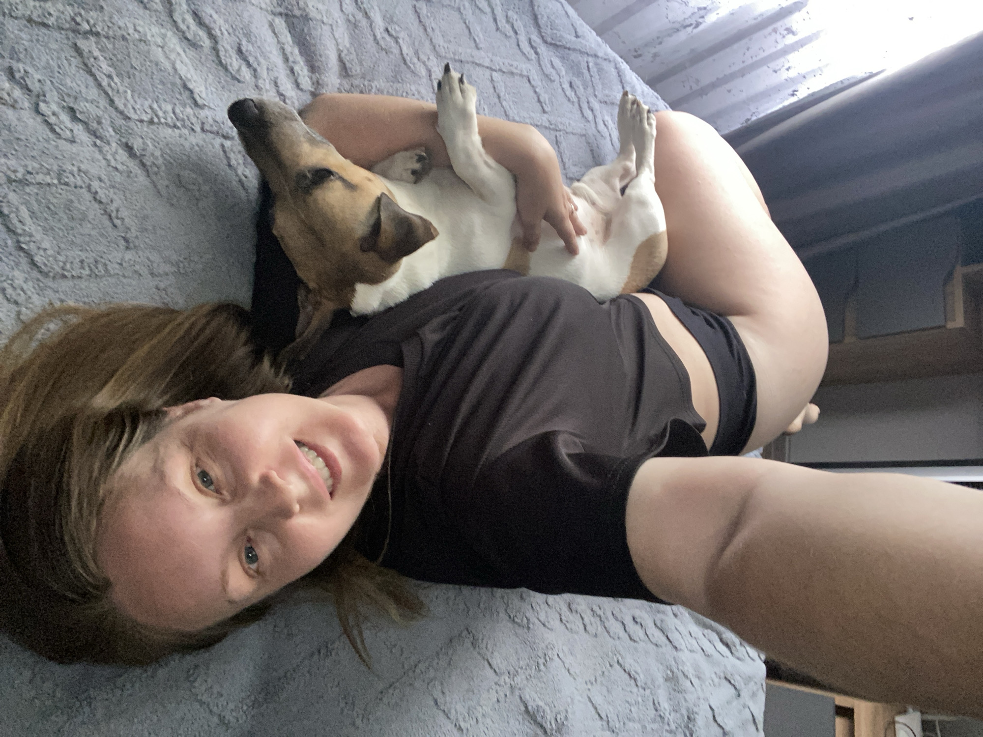 A woman lies on a gray textured surface, possibly a bed, with a dog lying on her chest. The woman is fair-skinned with brown hair and blue eyes, smiling at the camera. She wears a black shirt and black shorts. The dog is tan and white, lying on its back with its paws in the air. The background includes a window and a piece of furniture with shelves. The location, Noviy, Russia, cannot be determined from the image alone.