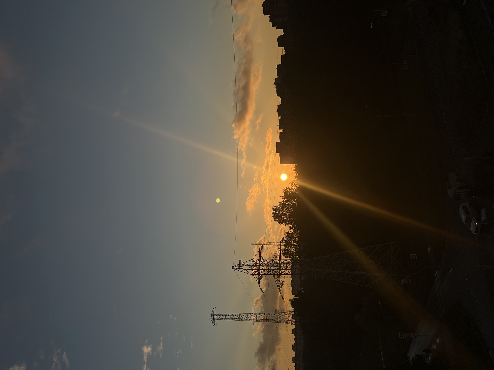 A low-angle, vertical shot captures a sunset over a residential area in Saint Petersburg, Russia. The sky transitions from a deep blue at the top to a warm orange near the horizon. Several high-voltage electricity pylons dominate the mid-ground, silhouetted against the vibrant sky. In the distance, the indistinct shapes of apartment buildings form a dark, horizontal line. The sun, a bright orange disc, is partially obscured by a power line, casting long sunbeams across the scene. Wispy clouds are scattered across the sky, tinged with orange from the setting sun. The lower part of the image shows a street scene in silhouette, with a few cars visible. The overall impression is one of urban twilight, with industrial infrastructure and residential buildings bathed in the dramatic light of sunset.