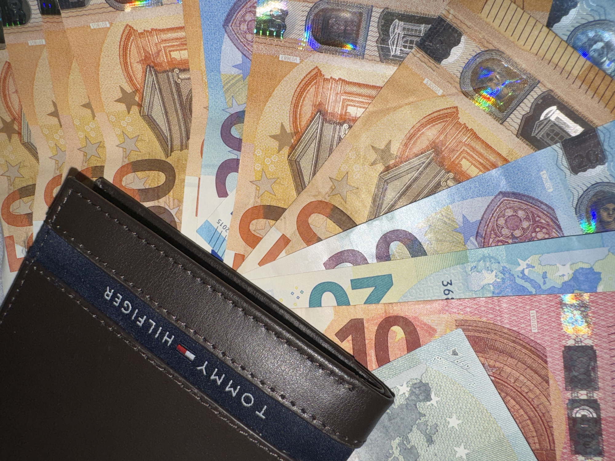 The image shows a brown Tommy Hilfiger wallet laying on top of a pile of euro banknotes. The banknotes visible are of various denominations, including 50, 20, and 10 euro notes. The notes are arranged in a semi-circular fan pattern. The location and city are not identifiable from the image.