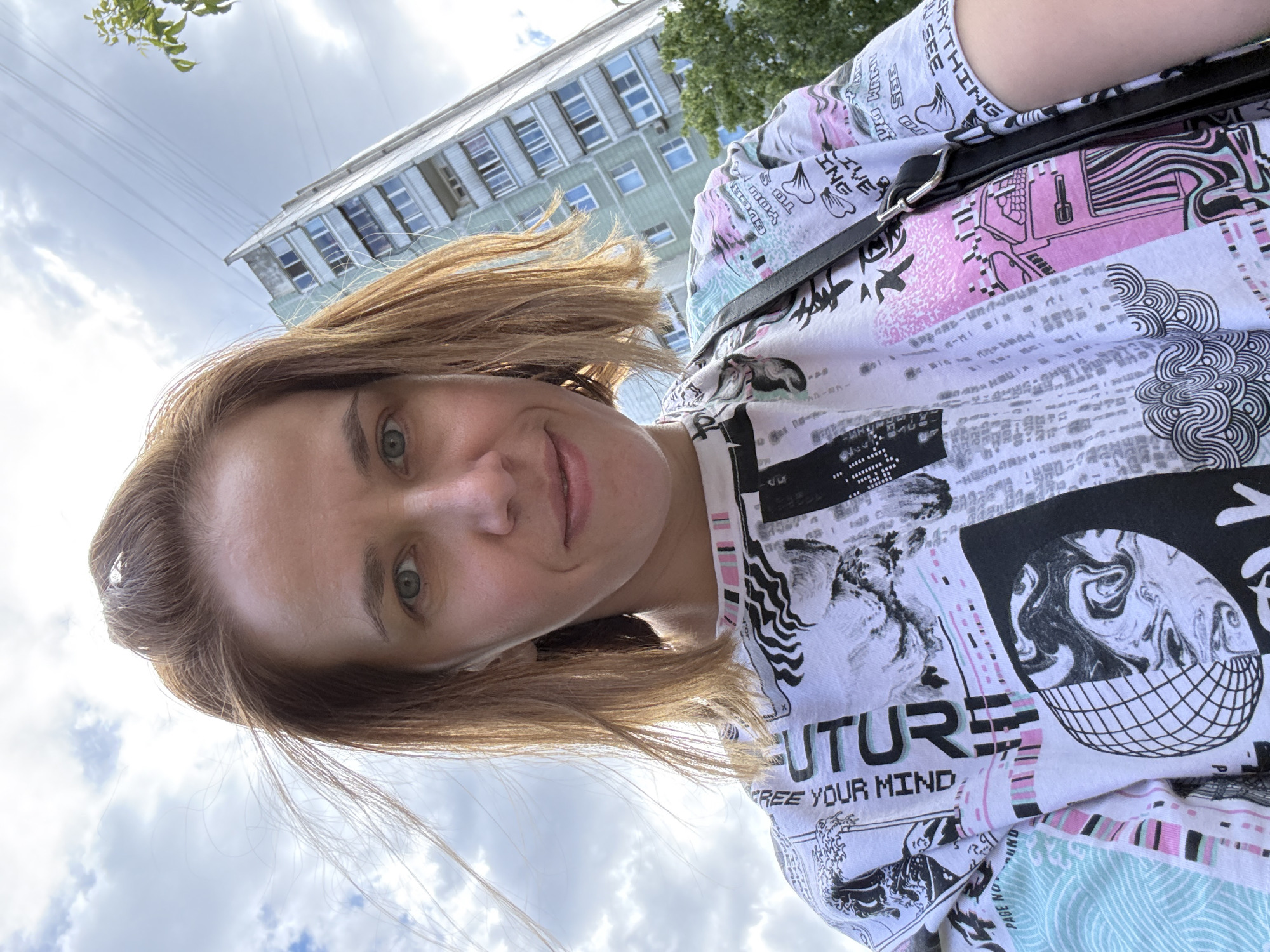 A medium shot shows a woman with short, light brown hair and green eyes. She is looking at the camera and smiling slightly. She is wearing a white t-shirt with a graphic print. A dark bag is slung over her shoulder. In the background, a tall, gray apartment building with many windows is visible against a cloudy sky. The building appears to be a typical Soviet-era residential block. The image is taken from a low angle, looking up towards the sky and the building. The presence of the apartment building and the style of its architecture are indicative of an urban environment in Moscow, Russia.