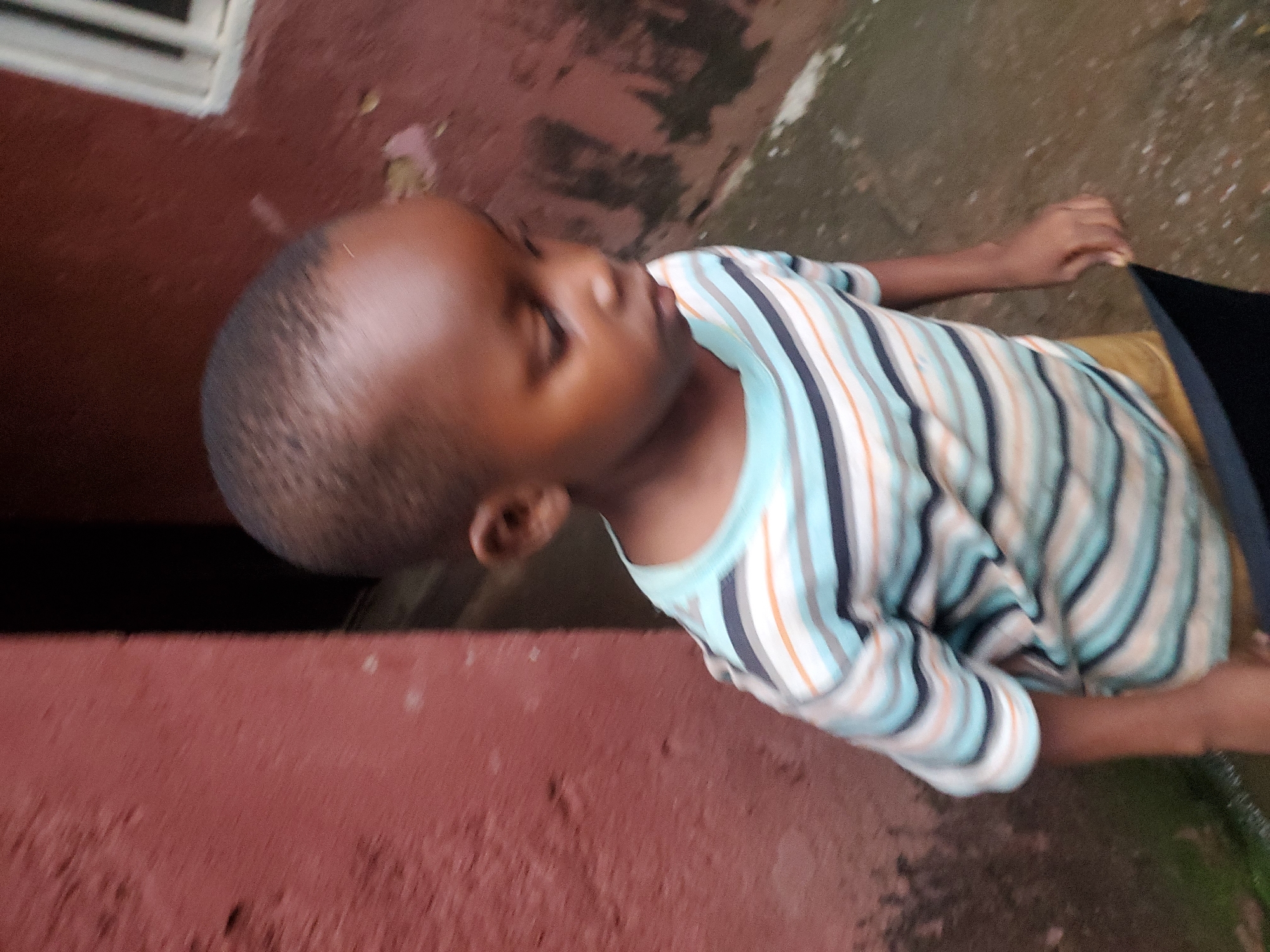 The image shows a close-up, tilted view of a young child wearing a striped t-shirt. The child's head is turned away from the camera, and their face is partially obscured, though they appear to be looking downwards. The t-shirt features horizontal stripes in light blue, dark blue, white, and orange. The background consists of a reddish-brown wall and a greyish, textured ground. A dark object, possibly a piece of cloth or bag, is visible to the right of the child. The lighting suggests it could be daytime, but the overall quality of the image, with a slight blur, makes it difficult to determine specific details about the weather or precise time of day. The setting appears to be outdoors, potentially in an urban or semi-urban environment in Jos, Nigeria, given the common textures of walls and ground seen in such locations. There is no visible text in the image.