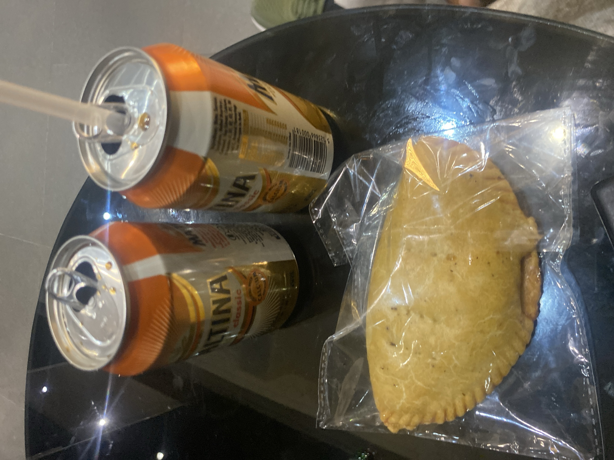 The image shows two open cans of Maltina Classic beverage and a pastry item in plastic wrap, all placed on a dark, reflective surface that appears to be a table. The cans have orange and silver labels with visible text. One can has a clear straw inserted into it. The pastry is golden brown and has a crimped edge. The lighting suggests an indoor setting, possibly a restaurant or home environment. The presence of the Maltina cans and pastry indicates a casual refreshment setting. The background is out of focus but shows hints of a tiled floor and possibly furniture. No individuals are visible in the image.