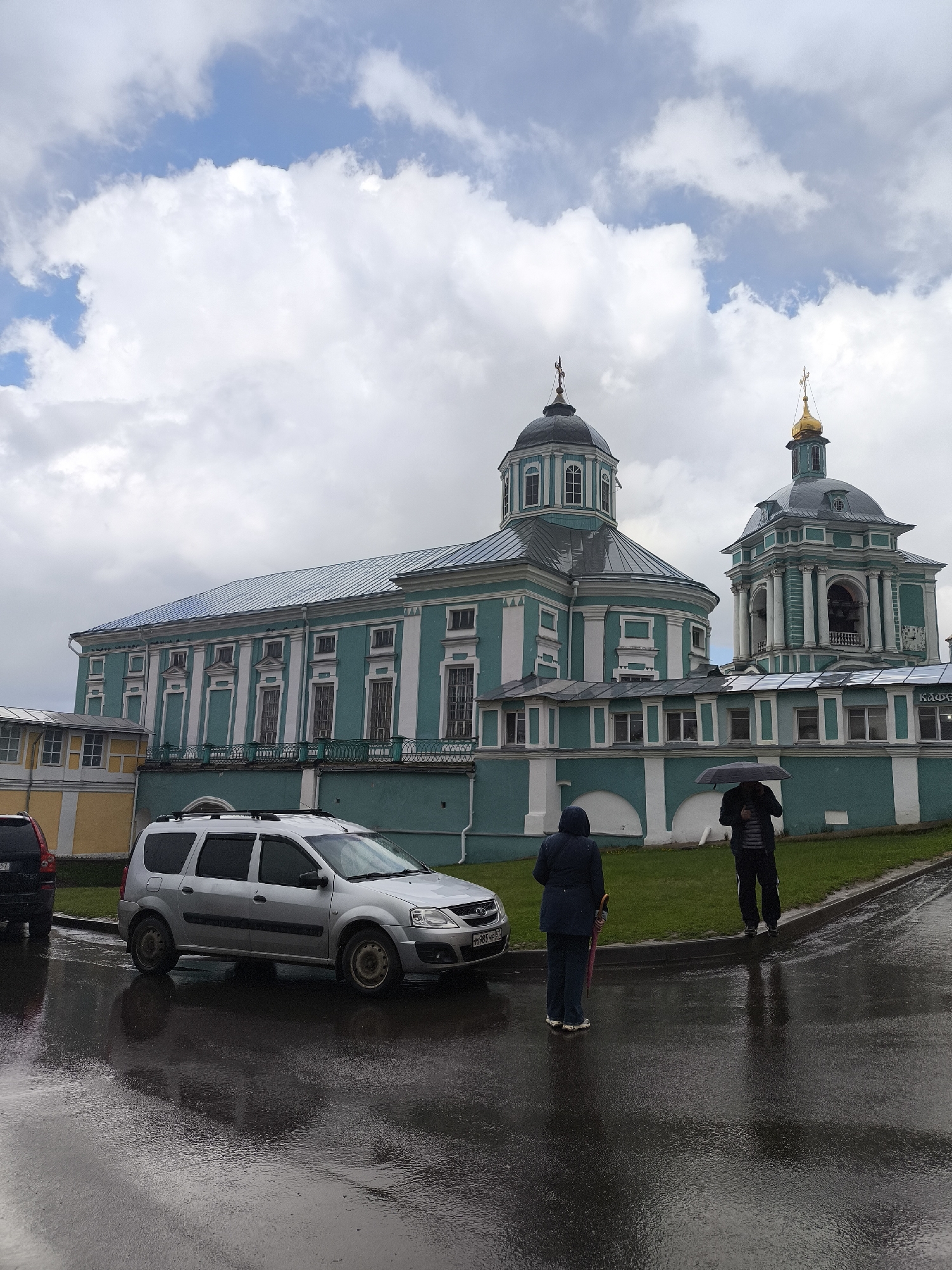 A street scene in Unknown, Unknown, on a rainy day, shows a large Eastern Orthodox church with turquoise and white walls and gold-domed towers dominating the background. The ground is wet, reflecting the sky and buildings. A silver Lada Largus car is parked on the left side of the road. Two people are standing on the sidewalk to the right of the car. One is facing away from the camera wearing a dark hooded coat and holding an umbrella. The other, also holding an umbrella, is wearing a dark jacket and dark pants. The sky is overcast with a mix of light and dark clouds.