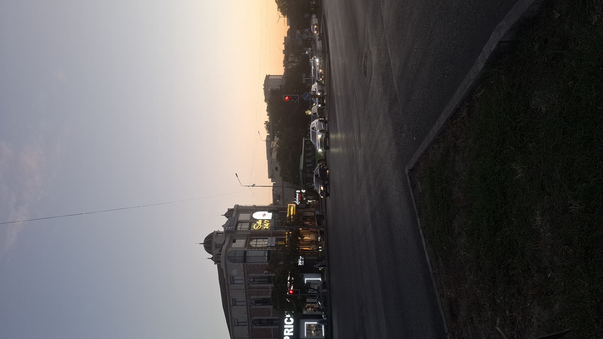 The image depicts a street scene in Shymkent, Kazakhstan, during what appears to be either sunrise or sunset, indicated by the soft, warm light in the sky. The foreground features a paved road with a patch of grass to the lower left, bordered by a concrete curb. In the mid-ground, several vehicles are visible on the road, including cars and a green bus, suggesting moderate traffic. A traffic light, red in color, is also prominent.
Dominating the left side of the image is a multi-story building with architectural elements that suggest it might be a commercial or residential complex. The building has large windows, and some of its facade is illuminated by signage. Notably, there is a sign that reads "SOHO 24/7" in illuminated letters. Another sign reads "PRICE" in large, white capital letters. People are seen walking on the sidewalk in front of the buildings, though their features are not distinct due to the lighting conditions.
In the background, more buildings and trees are visible, receding into the distance. The sky is a gradient of soft blue and pale orange, typical of twilight hours. A thin, dark line, possibly a cable or wire, extends diagonally across the sky from the upper left to the mid-right of the frame. The overall atmosphere is calm, with the urban environment bathed in the gentle light of either dawn or dusk.