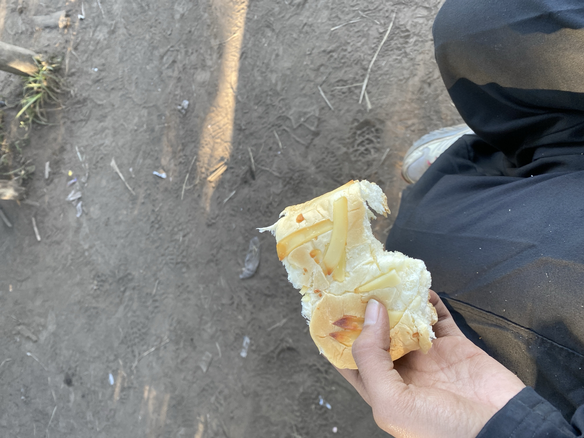 A person's hand holds a partially eaten bread roll filled with cheese and possibly some type of sauce, like ketchup or chili sauce. The person is wearing black pants and a white sneaker. The background appears to be an outdoor area with dirt and some dry plant debris, possibly in a rural or unpaved urban setting. The lighting suggests it is daytime, with shadows cast on the ground. The location is identified as Gondosuli, Indonesia.