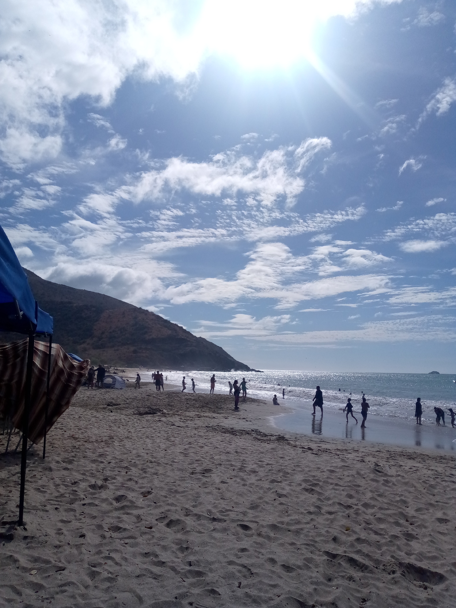 The image depicts a beach scene in Pedro González, Venezuela. The foreground is dominated by sandy beach with visible footprints. In the midground, numerous silhouetted figures of people are seen walking and playing along the shoreline. The ocean extends to the horizon under a bright, partly cloudy sky. A large, brown hill is located to the left of the beach. In the lower left corner, a portion of a beach umbrella is visible.