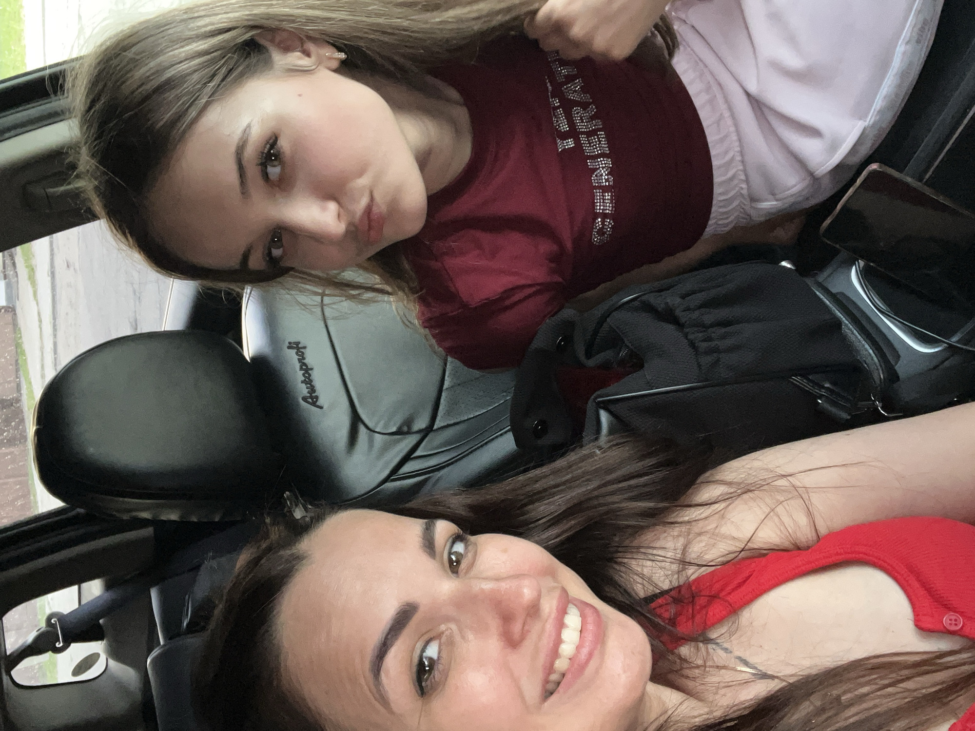 Two women are seated in the interior of a vehicle. The woman on the left, in the foreground, has long, dark brown hair and is smiling broadly at the camera, revealing her teeth. She is wearing a red top. The woman on the right is younger and has long, light brown hair. She is facing the camera with her lips pursed. She is wearing a dark red t-shirt with the words "GENERATION" in rhinestones and light pink pants. The interior of the car is visible, including black leather seats with the "Autoprofi" logo on the headrest. The background shows an outdoor scene with a road and buildings.