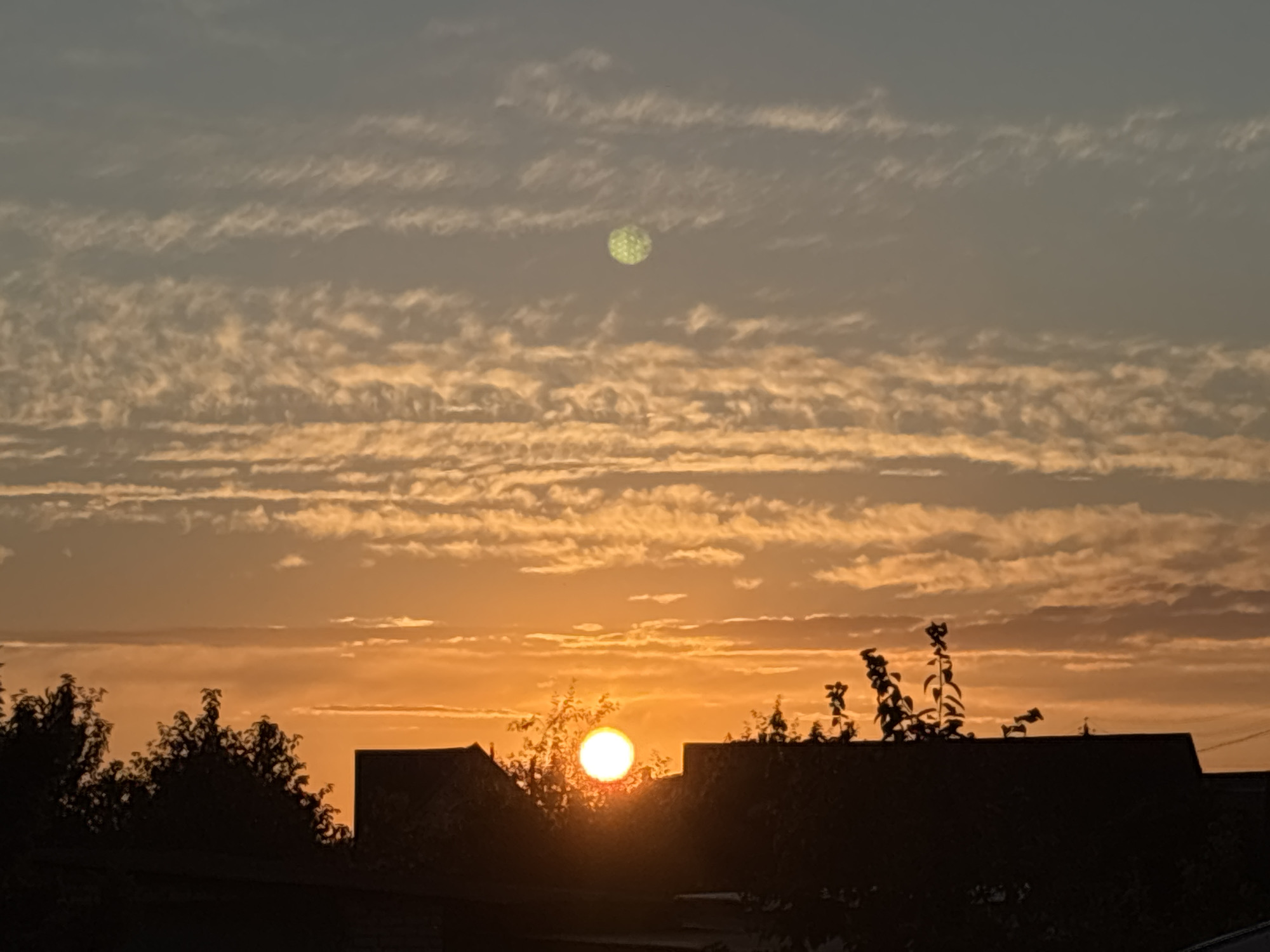 The image depicts a sunset over the city of Kapyĺ, Belarus. The sky is a vibrant canvas of orange and yellow hues, with scattered clouds forming horizontal bands. In the upper portion of the sky, a peculiar, bright green circular object with a textured surface is visible, possibly a lens flare or an atmospheric anomaly. Silhouetted against the glowing sunset are the rooftops of buildings and the dark outlines of trees, suggesting an urban or suburban setting. The sun itself is low on the horizon, casting a warm glow and partially obscured by the dark silhouettes in the foreground.