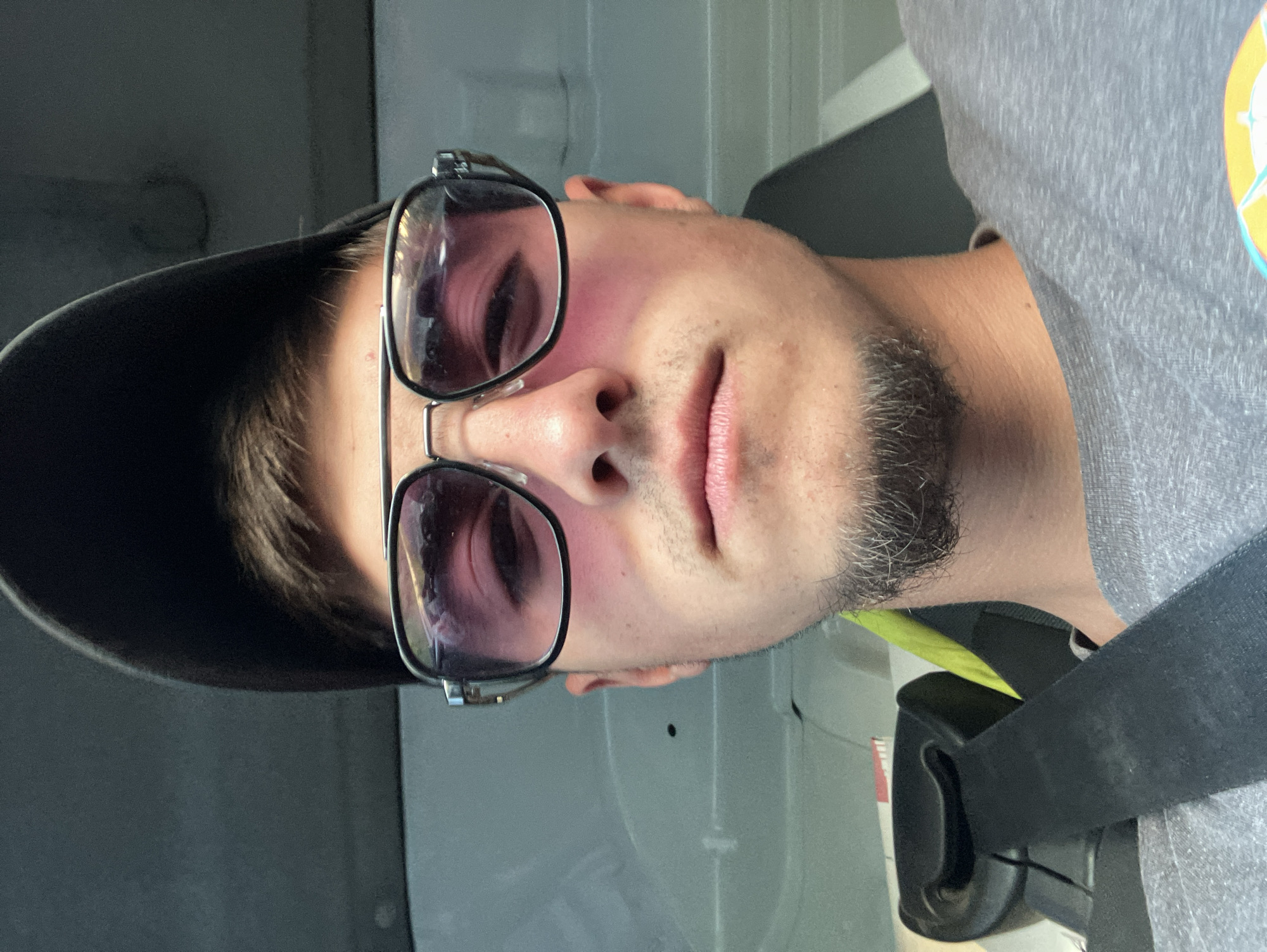 A close-up photo shows a man wearing sunglasses and a baseball cap in an indoor setting, possibly a vehicle. The man is wearing a gray shirt and a black seatbelt. Details about Komuniga, Bulgaria, cannot be determined from the image.