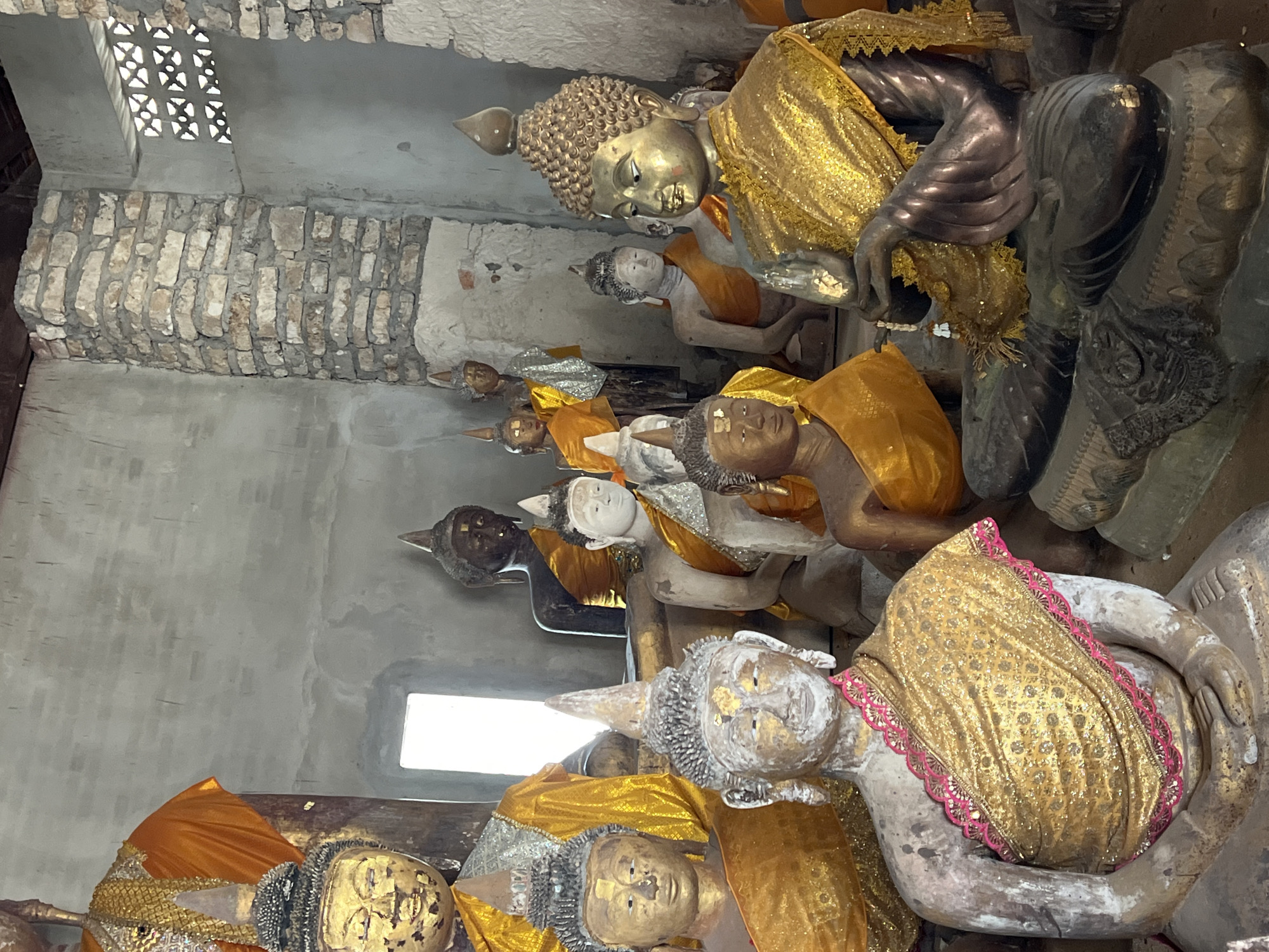 This image depicts a collection of Buddha statues, varying in size and material, housed within a rustic building. The statues are adorned with saffron-colored robes, some with intricate gold patterns. The architecture of the building appears to be a mix of concrete and exposed brick, with a window providing natural light. The overall setting suggests a temple or shrine in the city of Baan Harn, Thailand.