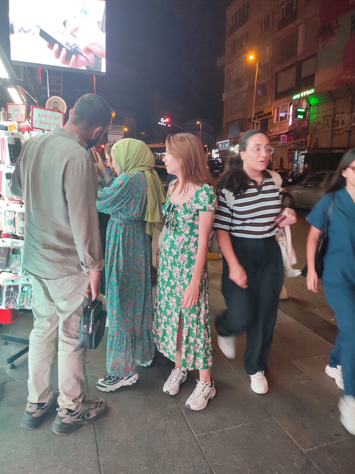 At night on a street in Kadıköy, Turkey, four individuals are visible. The person on the far left is a man wearing a muted green long-sleeved shirt and tan pants. He carries a black bag in his left hand. To his right is a woman wearing a light green hijab and a long, teal-colored dress with a floral pattern. Next to her is a woman with long, reddish-brown hair, wearing a green floral print dress. On the far right is a woman in a dark blue outfit. The individuals appear to be standing and walking on a paved sidewalk. In the background, a building with illuminated windows and storefronts with neon signs are visible. A large digital screen displaying an image is mounted above the storefront. A lamppost with a yellow light is also present.