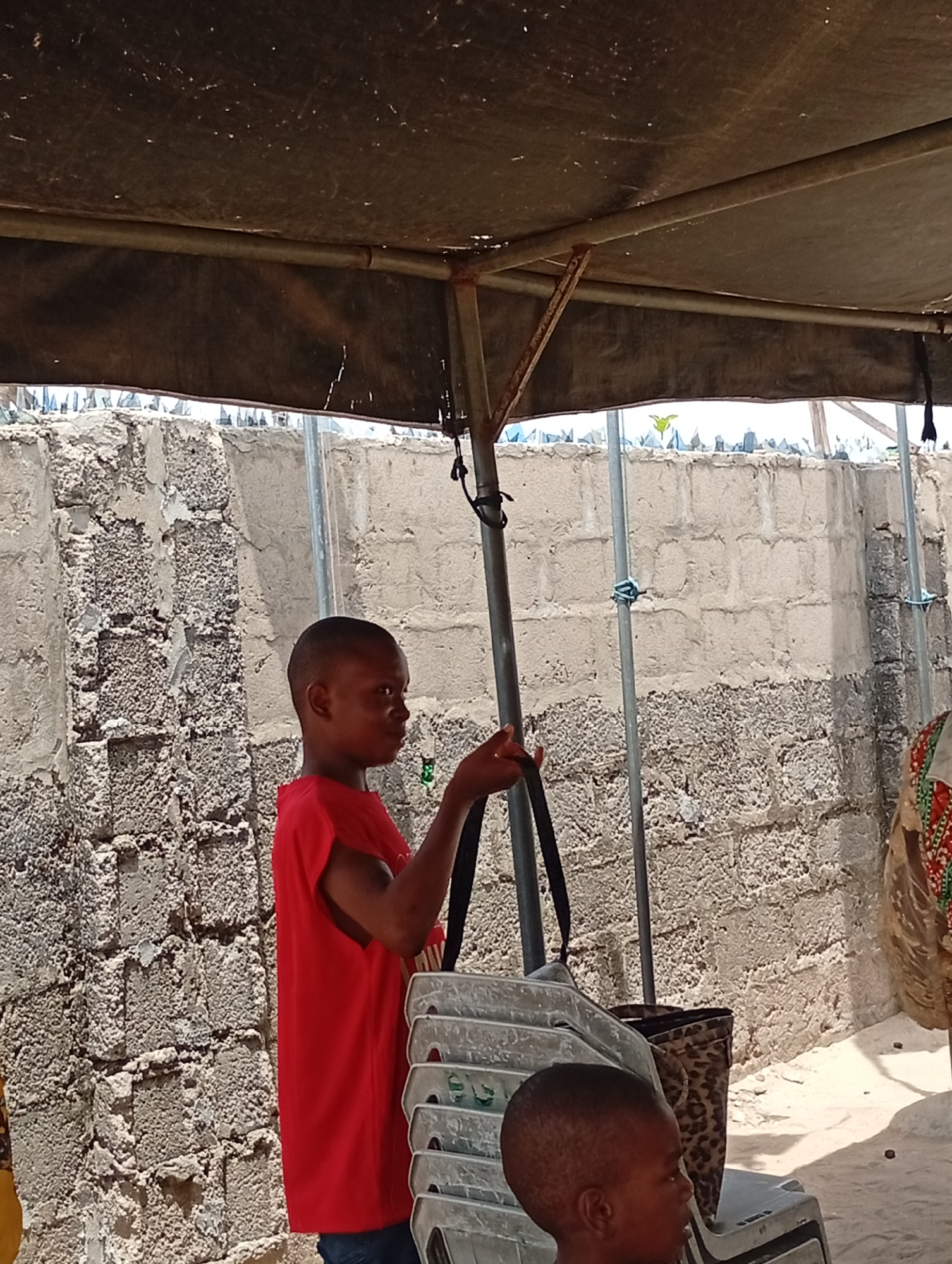 A young boy wearing a red shirt stands under a canopy in Adankolo, Nigeria. He's holding a bag with straps, and a stack of chairs are next to him. Another child's head is visible in the foreground. Behind them, a concrete block wall topped with what appears to be broken glass runs along the perimeter. The ground is sandy.