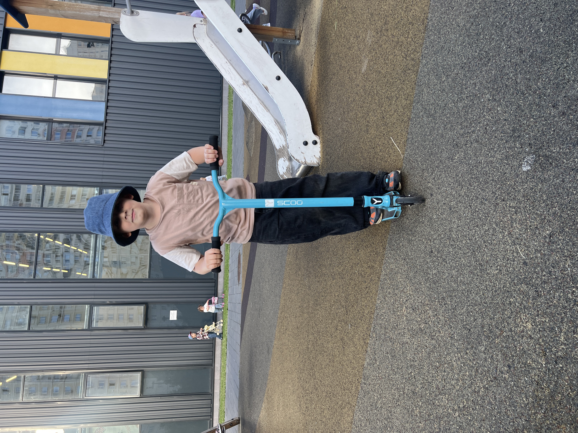 A child wearing a blue bucket hat and a light brown long-sleeved shirt is standing on a playground holding a blue scooter with the word "SCOO" printed on it. The child is wearing black pants and orange and black sneakers. The child is looking at the camera and smiling slightly.
The playground has a grey slide with white accents and a dark grey metal building in the background with colorful rectangular panels on the left side. The ground is covered in a soft, speckled material, with areas of light brown and dark grey. Two other children are visible in the background, one on a pink ride-on toy and another on a yellow toy. The scene appears to be outdoors during the day, with natural light. The overall mood is cheerful and playful.
The location is Mytishchi, Russia.