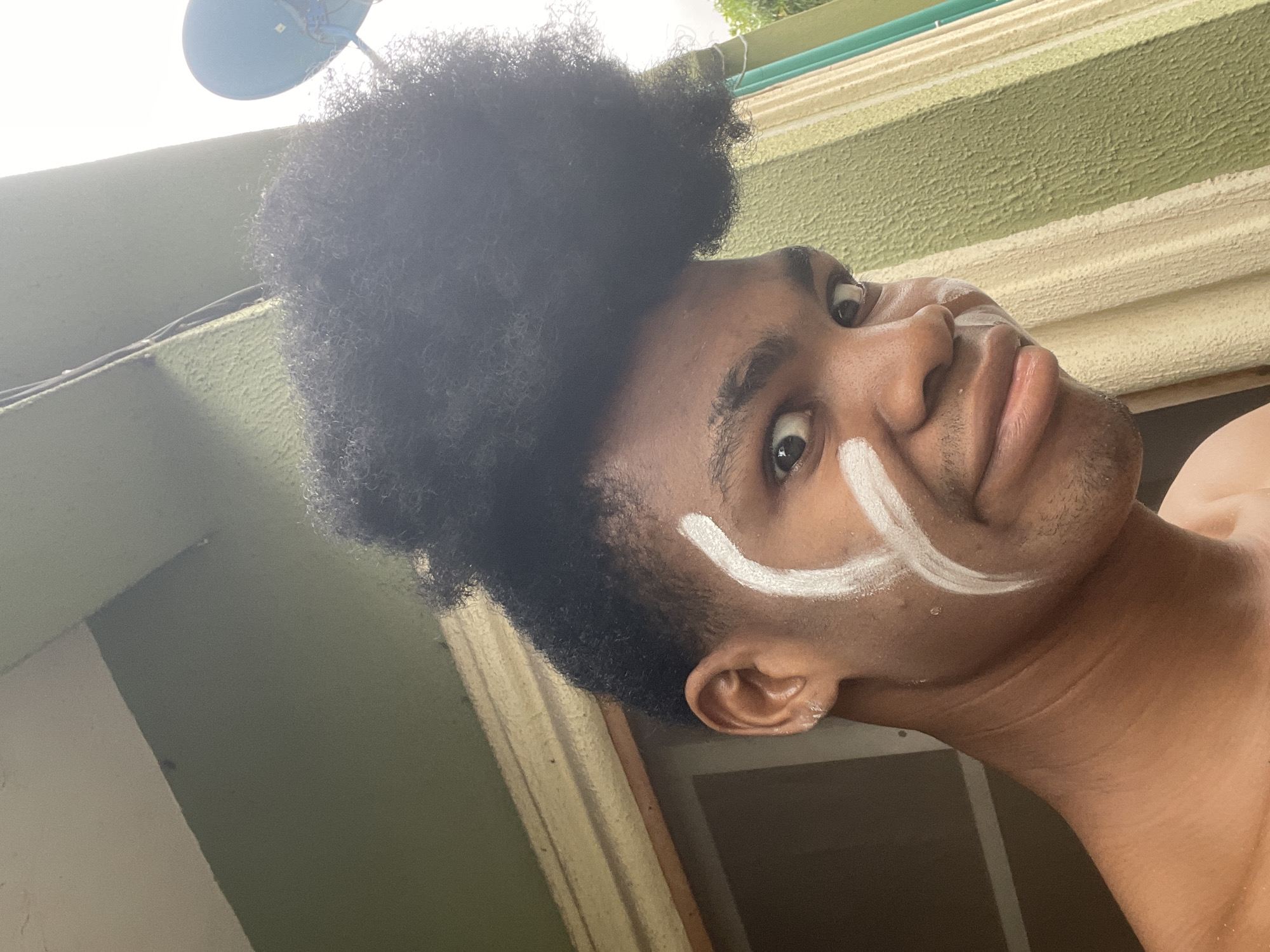 A low-angle, close-up shot depicts a person with dark skin and curly black hair. A white, curved line is painted on their right cheek. The background features a textured green and cream-colored wall. The person appears to be indoors, with natural light coming from above. Based on the instructions provided, the location is Ado, Nigeria, but the scene cannot be further specified. A satellite dish is visible in the top left corner. The person is shirtless.
