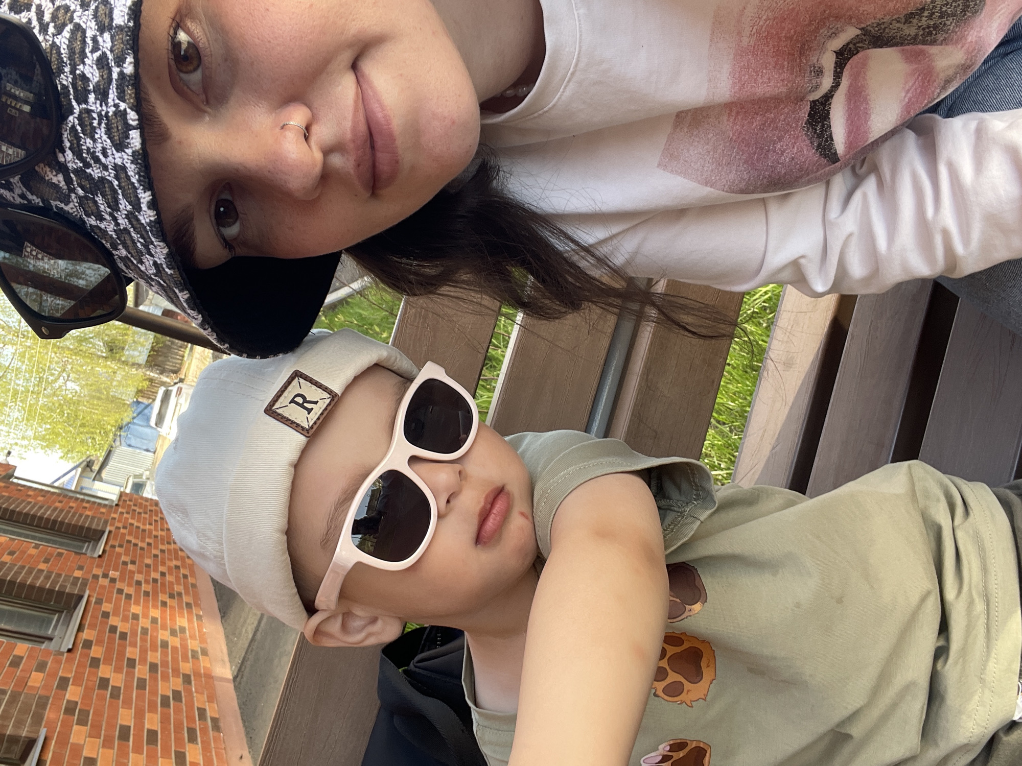 Two individuals are visible in an outdoor setting. A child wearing a beige beanie with a square patch bearing the letter "R" and sunglasses is lying down. An adult with a patterned hat and sunglasses perched on the hat is next to the child. They are on a wooden bench. A brick building is visible. The specific location of Izhevsk, Russia cannot be determined from the image.