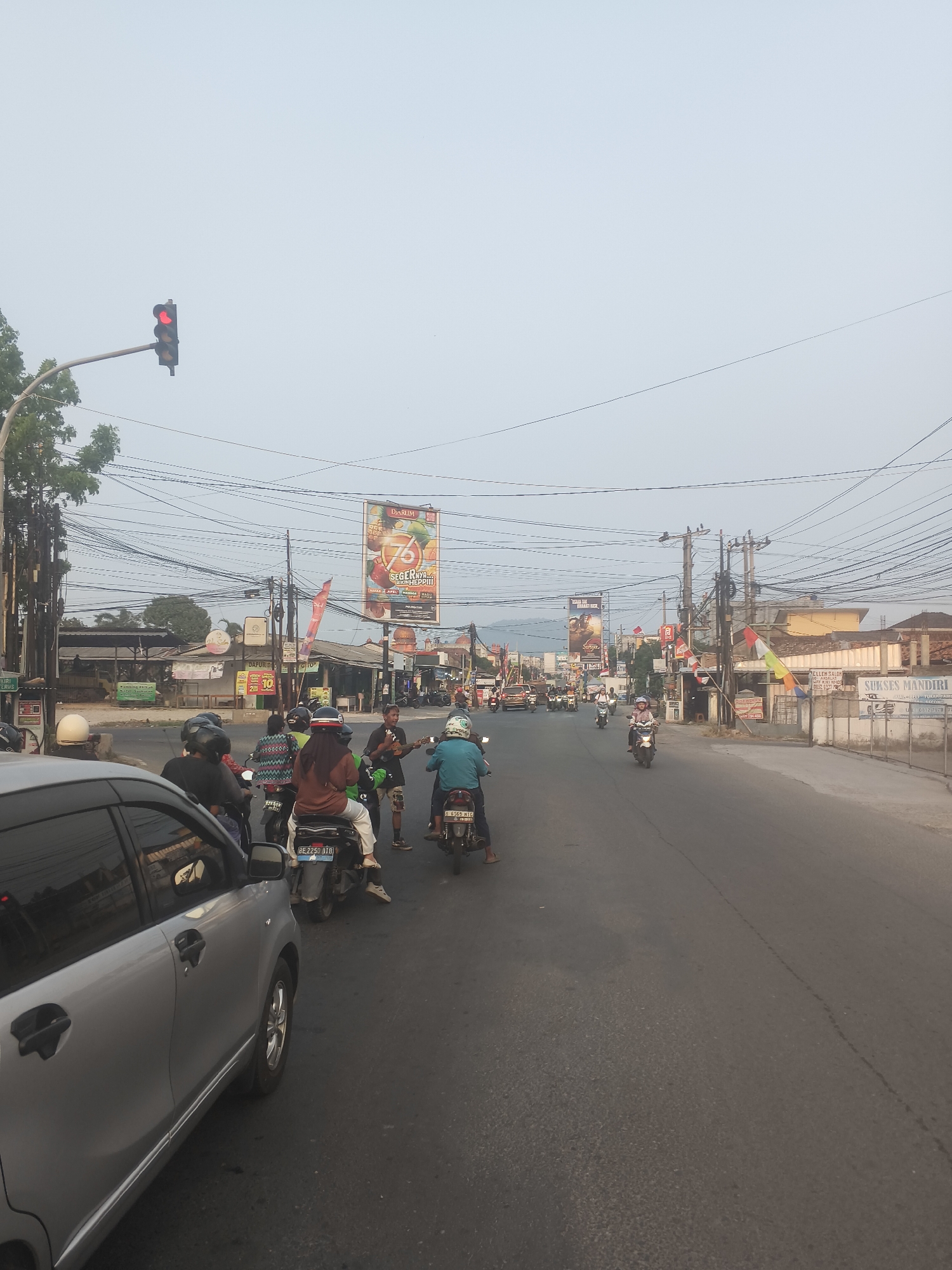 The image depicts a street scene in Bandar Lampung, Indonesia. A traffic light is visible in the upper left, showing a red signal. Multiple motorcycles are on the road, some with riders wearing helmets. A silver car is in the foreground on the left. In the background, there are buildings, overhead power lines, and billboards. One billboard prominently displays "76 SEGERNYA HEPIII" with images of fruits. Another billboard shows an advertisement for "DURUM". The sky is overcast.