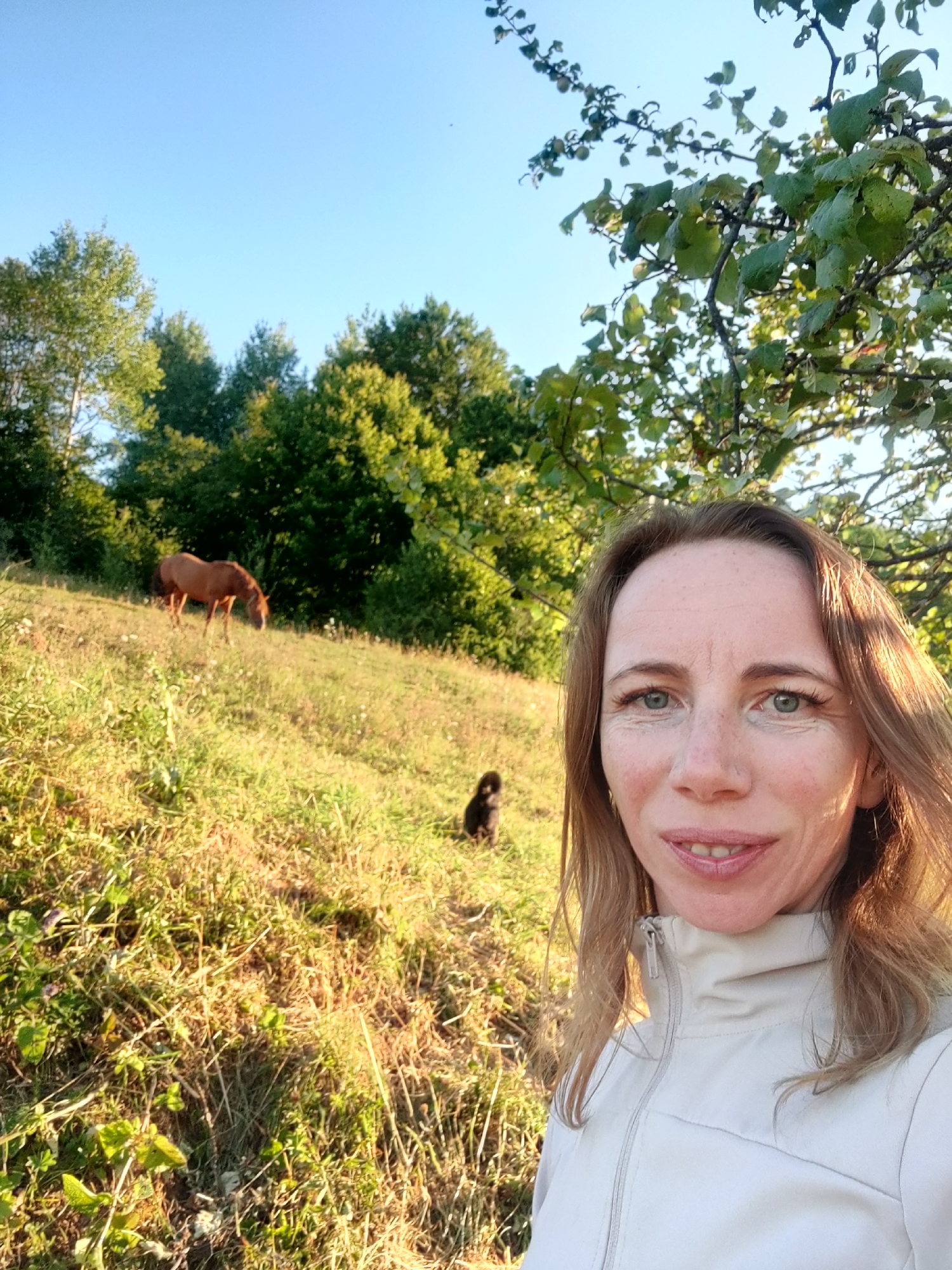 In Skotarske, Ukraine, a woman with light brown hair and blue eyes, wearing a light-colored jacket, smiles at the camera. Behind her, a brown horse grazes on a grassy hillside bathed in the warm light of a sunny day. A small black dog is visible further up the hill, near the horse. The background consists of lush green trees and a clear blue sky.