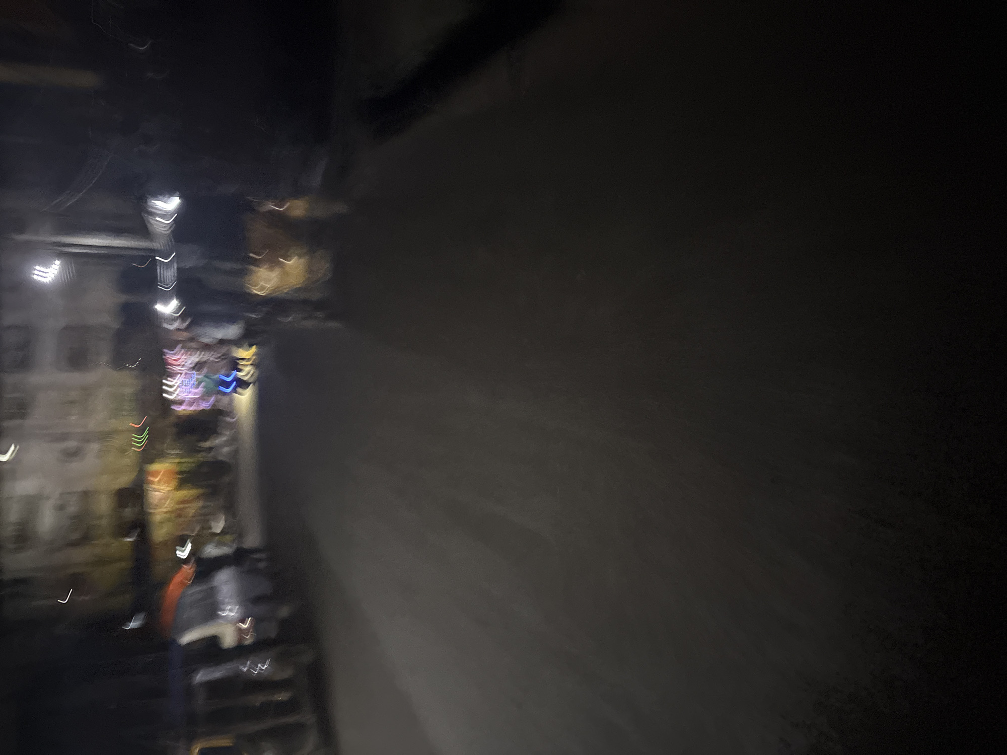 The image depicts a blurry, nighttime street scene in Onitsha, Nigeria. A street is visible with vehicles on the left side, possibly parked or moving slowly. The background shows buildings, some with illuminated windows or signs, suggesting an urban environment. Several figures, indistinct due to motion blur, are present on the street and sidewalk. The overall impression is that of a busy, active street at night, with artificial lighting creating streaks and halos due to the low light conditions and camera movement.