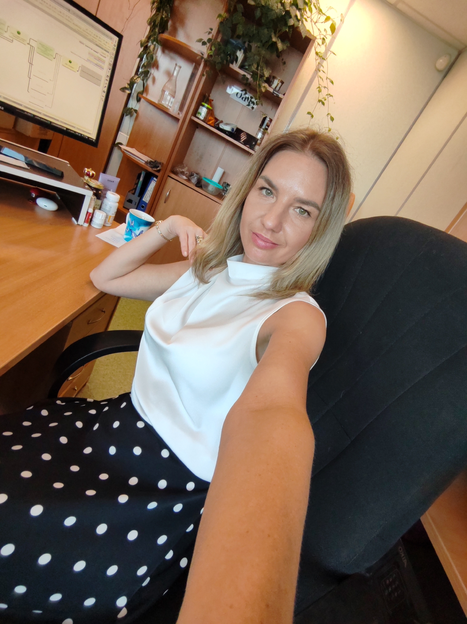 A blonde woman in a white sleeveless top and a black polka dot skirt is sitting in a black office chair, taking a selfie. She is seated at a wooden desk in what appears to be an office. On the desk, there is a computer monitor displaying a flowchart, a stack of papers, a blue mug with a butterfly design, and several bottles of pills. Behind her, there is a wooden bookshelf with decorative plants and various office supplies. The background wall is light-colored.