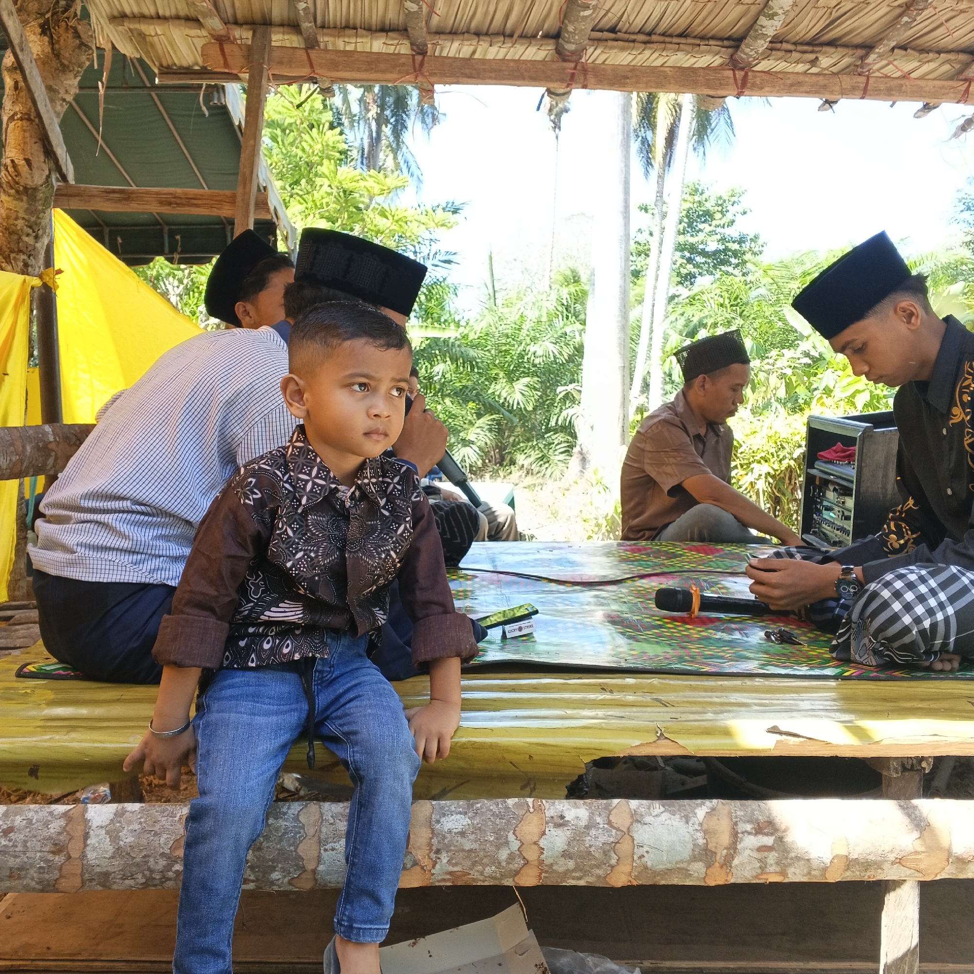 In Aluekeutapang, Indonesia, a group of people are gathered on a raised wooden platform, partially covered by a thatched roof. In the foreground, a young boy, wearing a patterned brown shirt and blue jeans, sits facing left, looking away from the camera with a contemplative expression. He wears a silver bracelet on his left wrist. To his left, partially visible, is a man in a light-colored, striped long-sleeved shirt and dark pants, also wearing a black peci (cap). Behind the boy, another man in a brown short-sleeved shirt and grey pants is seated, looking down at something in front of him, possibly interacting with a sound system. To the right, a young man in a black long-sleeved shirt with gold embellishments and a black peci is seated, looking intently at his hands, which are resting on a small electronic device, possibly a phone. He wears checkered pants. A black speaker box with red fabric inside is positioned to his right. The platform is covered with a colorful, patterned mat, on which rests a black microphone with an orange pop filter. A packet of "Lucky Strike" cigarettes is also visible on the mat. The background reveals lush green vegetation and palm trees, indicating a tropical environment. A bright yellow tarp is visible on the far left, attached to a wooden support. The overall scene suggests an informal gathering or event taking place outdoors.