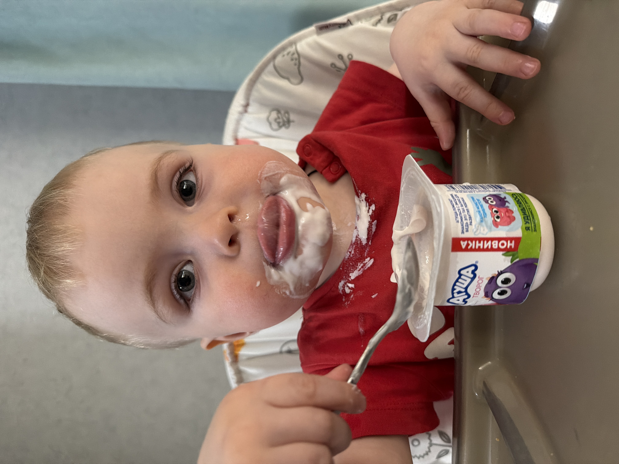 A young child sits in a highchair, facing the camera. The child has fair skin, light hair, and gray eyes. The child's face and red shirt are covered in white food, possibly yogurt. The child's tongue is extended with a blob of yogurt on it. In the child's hand is a spoon also covered in yogurt. Next to the child is a container of Ausha brand yogurt, which has cartoon characters on the container. The highchair is white with a gray tray. The background is a light blue wall. The location is likely indoors in Russia given that "Ausha" yogurt is a Russian brand. It's not possible to ascertain that the location is in Votkinsk.