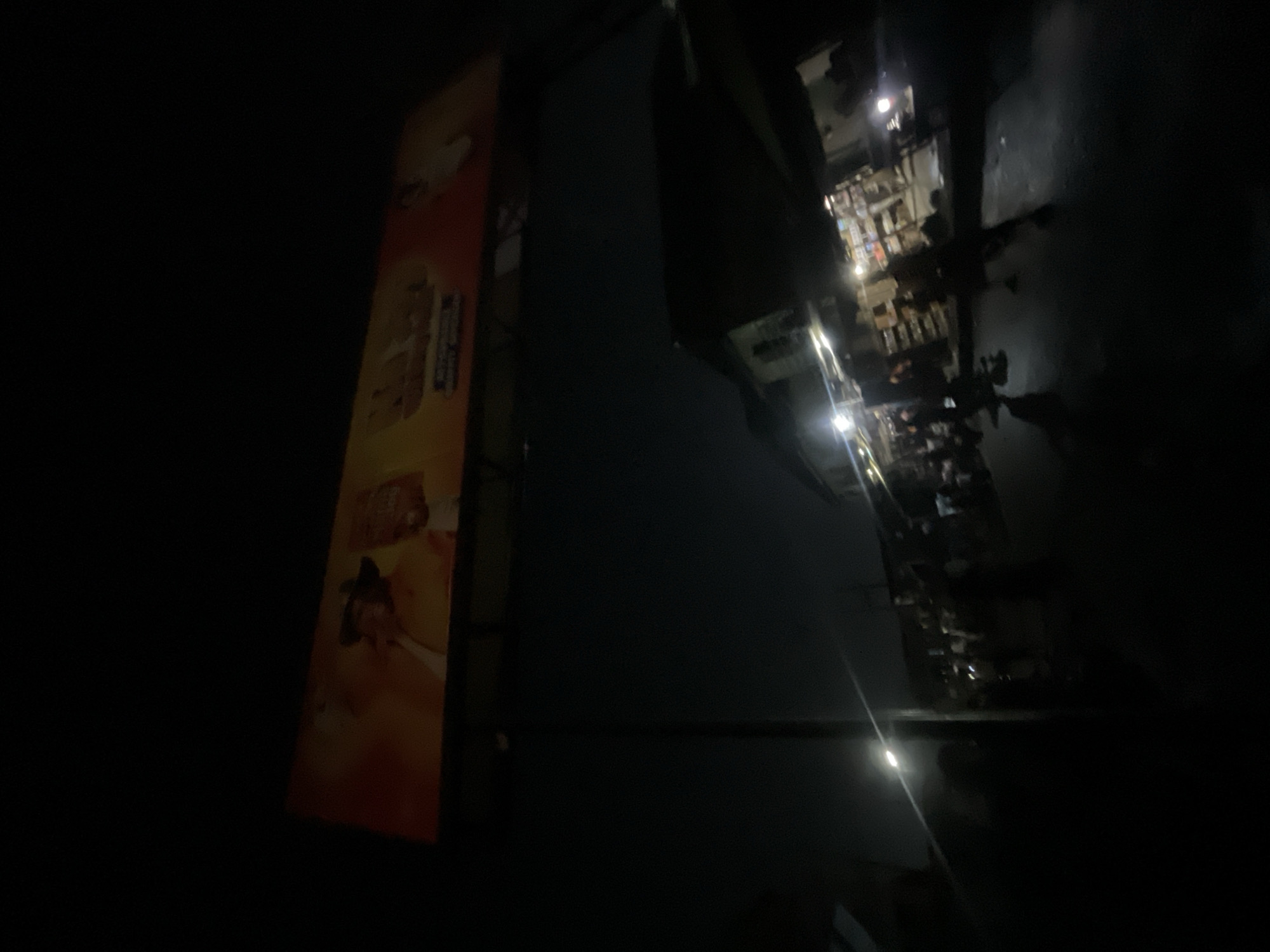 In Akowonjo, Nigeria, an outdoor billboard displaying a smiling individual wearing a hat and yellow clothing is illuminated against a dark night sky. Below the billboard, a street scene unfolds with a dimly lit market or shops. Figures of people are visible, some appearing to be browsing or walking through the area. The environment is characterized by artificial lighting from the shops and streetlights, casting reflections on the wet or dark asphalt of the street. The overall atmosphere suggests a bustling nighttime environment in a Nigerian city.