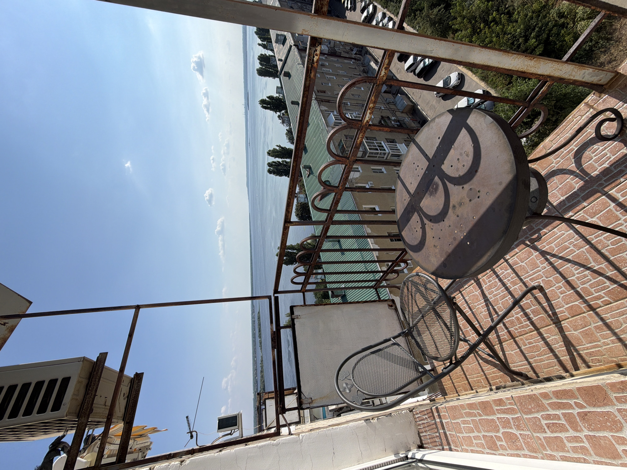 The image shows a view from a balcony overlooking a wide river in Saratov, Russia. The balcony has a metal chair and a small, round metal table with a decorative design on its surface. The floor of the balcony is tiled with a pattern of reddish-brown bricks. In the background, across the river, is a cityscape with several apartment buildings and trees. The sky is blue with a few white clouds. The scene suggests a warm, sunny day.