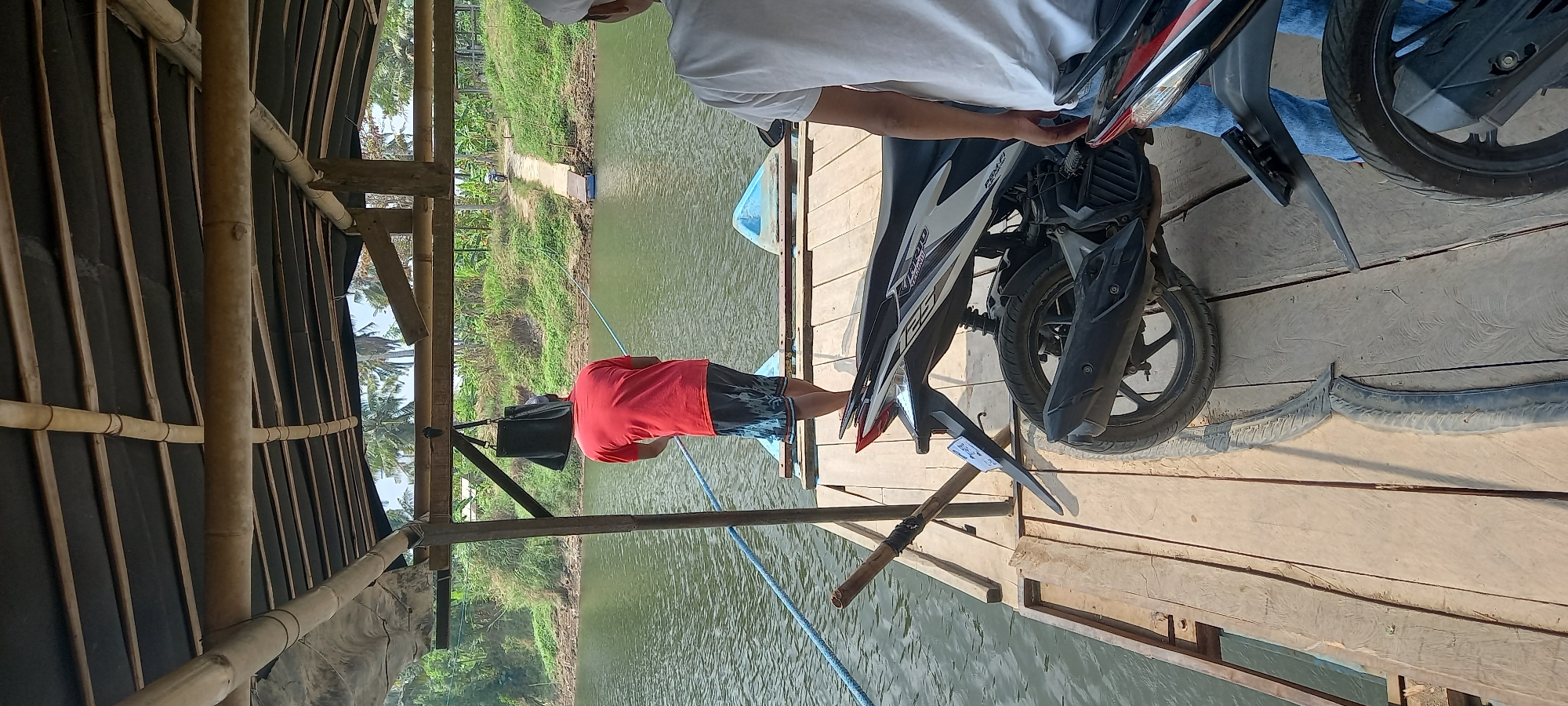 A man in a red shirt and patterned shorts stands on a wooden pier, facing away from the camera and holding a fishing rod. He is positioned under a bamboo-covered shelter, with a dark bag hanging from a support beam. The pier extends over a calm, green body of water. To the right, two motorcycles are parked on the pier. In the background, lush green vegetation and palm trees line the opposite bank of the water. A narrow dirt path leads up from the water to a higher elevation. Two other individuals are partially visible on the pier; one on the far right wears a white hat and shirt, and another person's legs in blue jeans are visible near the motorcycles. The scene appears to be a quiet, rural location in Cintaratu, Indonesia.
