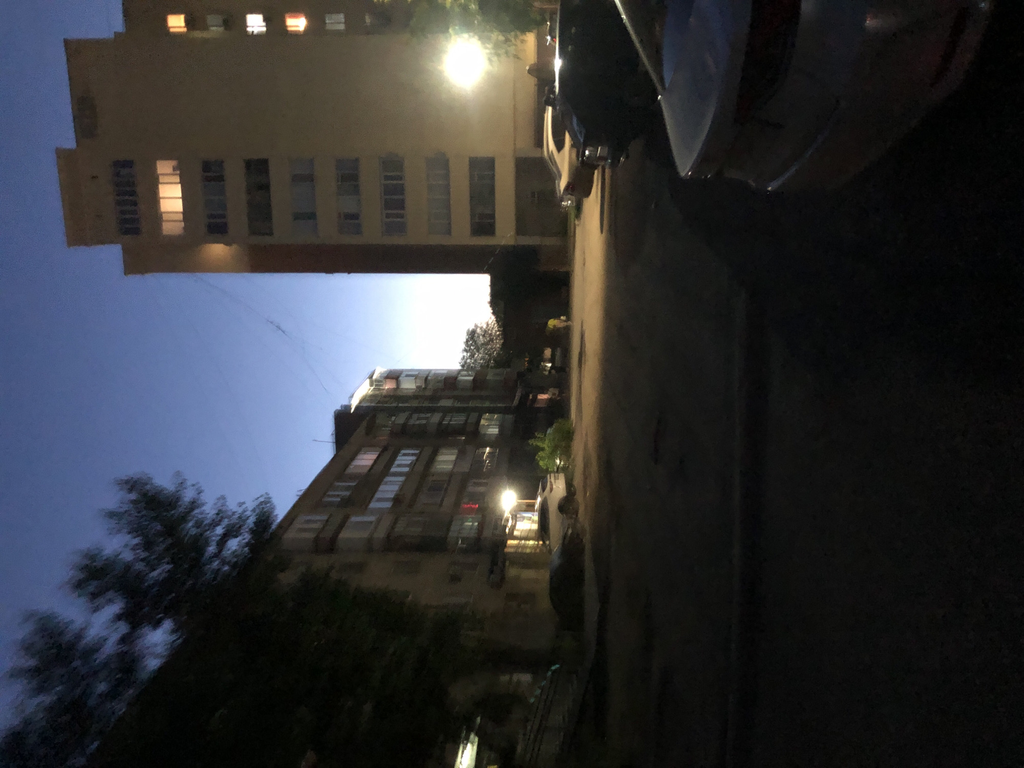 The image depicts an outdoor scene at dusk in an urban environment. A tall, multi-story building with lit windows stands on the right side of the frame, casting a warm glow against the twilight sky. To the left, another apartment building is visible, also with illuminated windows. The ground in the foreground appears to be a paved area, possibly a courtyard or parking lot, with several cars parked. One white car is prominently parked near the center, with its headlights on. The scene is dimly lit, suggesting the time of day is either early morning or late evening. Trees are present, with foliage visible on the left, and their silhouettes are dark against the sky. The overall impression is of a residential area at the end of the day.