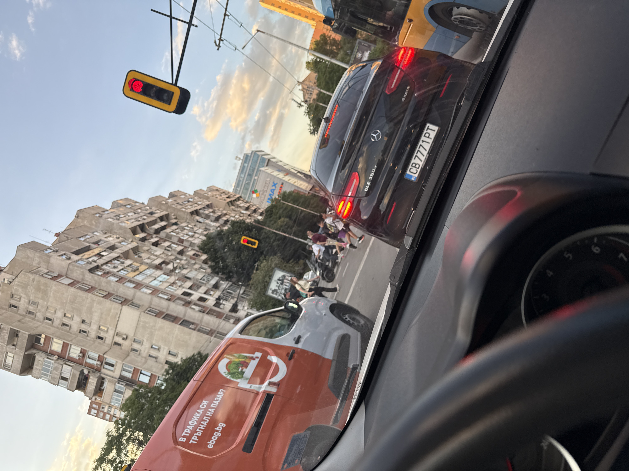 The image depicts a street scene in Sofia, Bulgaria, taken from the interior of a car. The car is stopped at a red traffic light. In front of the car, there is a Mercedes GLE 350d with a license plate CB 7771 PT and an ebag.bg delivery van. Pedestrians and a scooter are crossing the street. In the background, there are high-rise buildings, including one with the "IMAX" logo. The sky is partly cloudy.