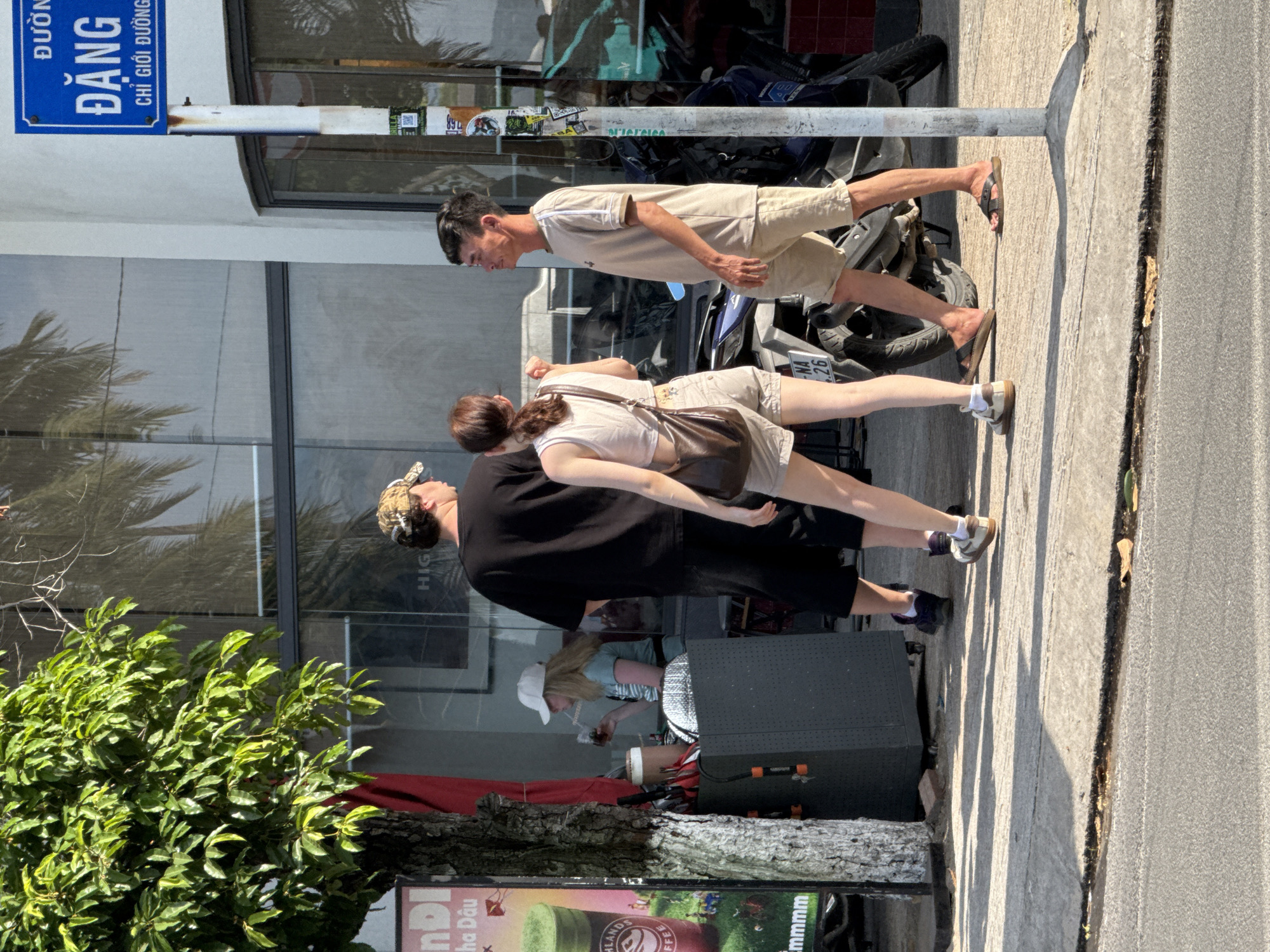 In Bac Nha Trang Ward, Vietnam, three individuals are present on a sunny day along a sidewalk. A woman in a white cap and light blue shirt is seated at a small outdoor counter, drinking from a straw. Two men and one woman are standing and walking on the sidewalk. The man in the foreground on the right wears a tan t-shirt and shorts, walking with a slight lean. Next to him, a woman in a light-colored crop top and tan shorts walks away from the camera, carrying a brown shoulder bag. A man in a black t-shirt and shorts stands to the left, his back partially to the camera, wearing a camouflage baseball cap. Behind them, a motorcycle is parked. The environment includes a building with reflective windows, a leafy tree on the left, and a street sign in the upper right corner written in Vietnamese. The sidewalk is concrete, with shadows cast by the tree and people.