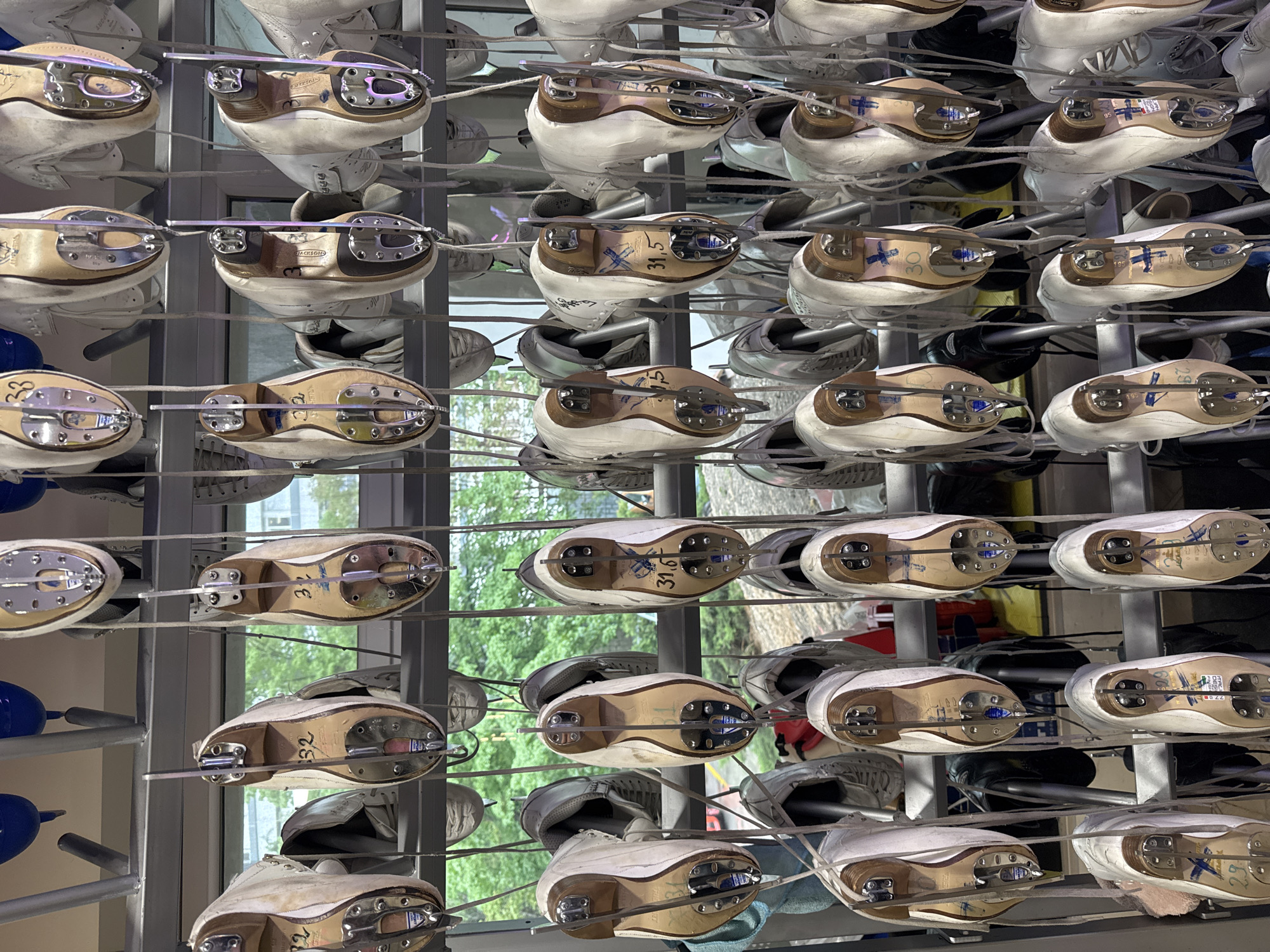 The image depicts a metal shelving unit densely packed with ice skates, presumably in a rental facility or storage area. The skates are predominantly white with brown soles and silver blades. They are arranged in rows and columns, suggesting an organized system for storage or display. The blades of the skates are secured by metal attachments. Some skates have handwritten numbers on the soles, possibly indicating the size of the boot or an inventory number. Through the window in the background, a glimpse of greenery, possibly trees, is visible, and the exterior architecture of a building can also be seen. Given the city of Tbilisi, Georgia, as context, the image could be taken within a skating rink or a sports complex. No people are visible.