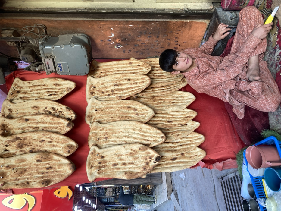 The image portrays a street scene in Quetta City Tehsil, Pakistan. A young male, estimated to be between 10 and 14 years old, sits cross-legged on a colorful patterned rug placed over a maroon cushion. He wears a light brown or peach-colored, pleated traditional shalwar kameez, with his bare feet visible. He holds a yellow-handled utility knife or tool in his right hand, resting it on the rug, and looks directly at the viewer with a neutral expression.
Behind him, to his left, is a vertical display of approximately 20-25 oven-baked flatbreads, resembling naan, arranged in two overlapping rows on a red fabric background. These flatbreads are oblong, exhibiting typical blistered and charred surfaces from baking. To the immediate right of the bread display, a grey gas meter is mounted on a weathered reddish-brown wall.
In the lower-left foreground, adjacent to the seated boy, a blue plastic basket contains several small metal bowls, a plastic jug, and other miscellaneous containers. In the background, to the left of the bread display, an open storefront is visible, featuring red signage with yellow Arabic script. Inside this store, a male individual wearing a dark shirt and light-colored pants is partially visible, appearing to be attending to items within the establishment. The ground surface in the foreground and extending into the background is concrete pavement, with a metal grate visible on the far left.