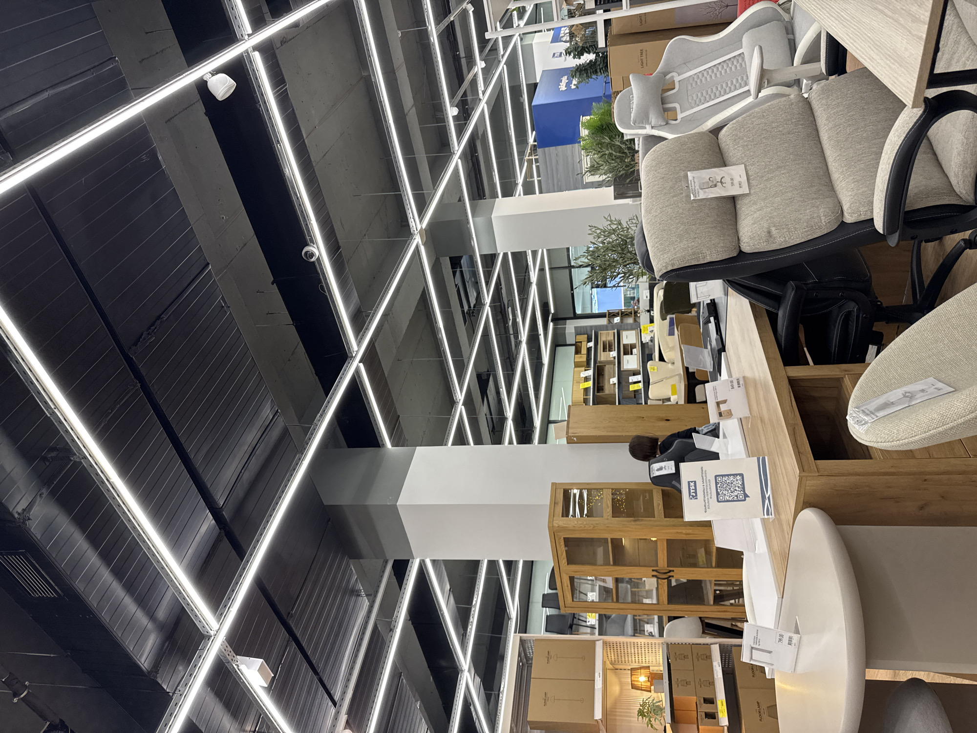 The image captures the interior of a retail store, likely a furniture or home goods store. The location is in Tbilisi, Georgia, as indicated by the QR code on the card reading "JYSK Грузинский магазин, Тбилиси".
The focus is on a display area featuring several office chairs in various styles and colors (grey, white), and wooden desks and cabinets. There is also a person, with dark hair, at the back behind a desk.
The environment has a distinct industrial-modern aesthetic. The ceiling is dark and crossed with a grid of bright, linear lighting fixtures.