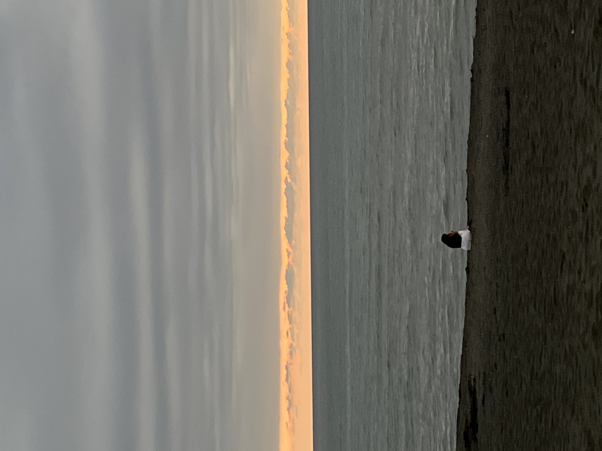 A solitary person, viewed from behind, sits on a dark sandy shore, facing a vast, gray ocean under an overcast sky. A thin band of pale orange light, indicative of sunset or sunrise, is visible on the horizon, separating the sea from the clouds. The water exhibits moderate waves. The individual is wearing a white garment and has dark hair. The environment suggests a coastal setting in Ureki, Georgia, known for its beaches. The overall atmosphere is serene and contemplative.