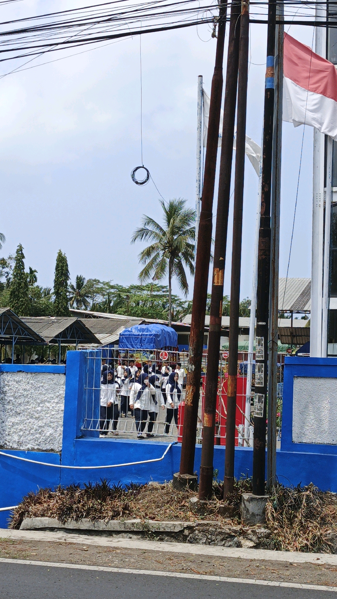 The image depicts a street scene in Sukaresik, Indonesia. In the foreground, a paved road runs horizontally. Adjacent to the road is a low concrete ledge covered in dry vegetation and some scattered debris. Behind this ledge, a bright blue wall forms the lower boundary of a property. Above the blue wall, a metal fence encloses an area where a group of people, predominantly young individuals in white uniforms and dark headscarves, are standing in formation. They appear to be participating in an outdoor gathering or event. Several utility poles, some appearing weathered and rusty, stand in the mid-ground, with a tangle of overhead electrical wires crisscrossing the sky. A red and white flag, likely the Indonesian flag, is visible flying from one of the poles. In the background, a palm tree stands tall against a bright, overcast sky, with the roofs of several buildings and more trees visible further back. The overall impression is a typical daytime scene in a moderately urban or semi-rural area of Indonesia.