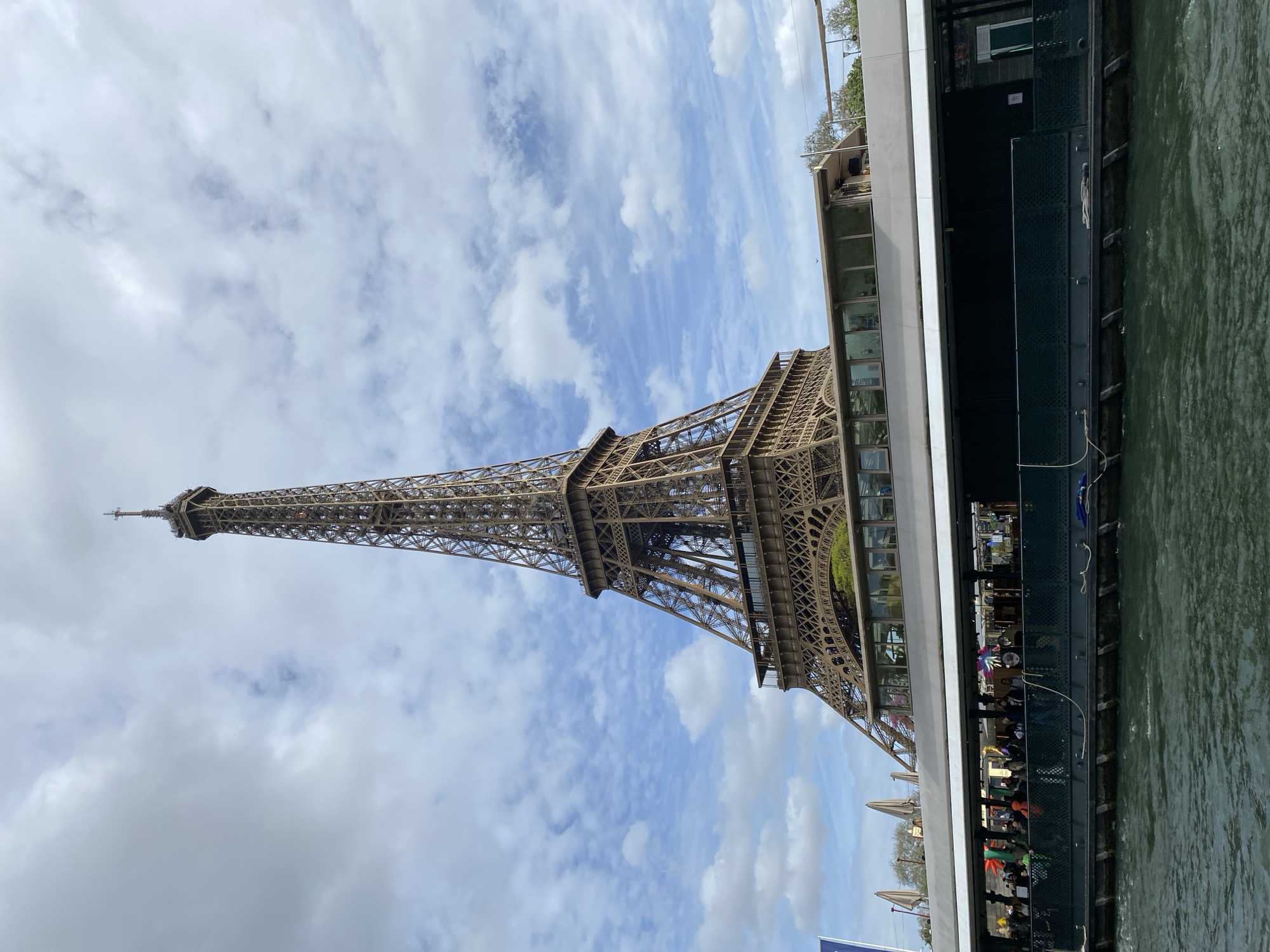 The image shows the Eiffel Tower in Paris, France, partially obscured by a boat. The Eiffel Tower is visible against a backdrop of a blue sky with scattered white clouds. The boat occupies the right side of the frame and appears to be a river cruise or tour boat with people seated inside. The water is visible beneath the boat.