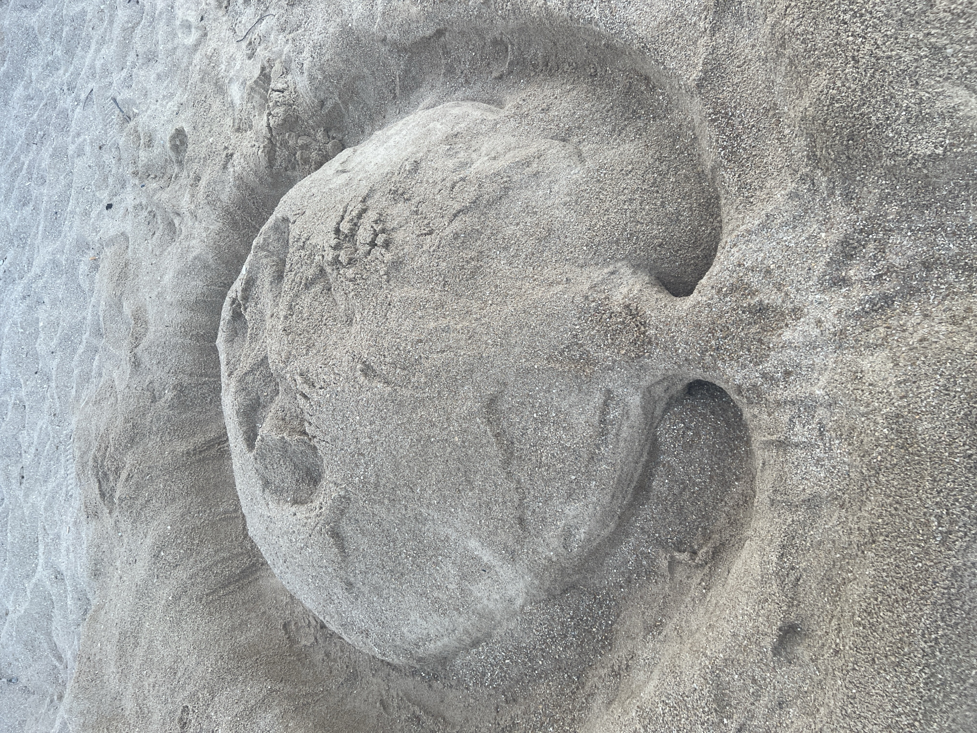 A sand sculpture resembling a face is situated on a sandy beach. The sculpture features a round head with indentations for eyes and a mouth. The surrounding area is composed of sand, with ripples and textures indicating a beach environment. The scene is captured from a high-angle perspective, focusing on the sand artwork. The location is identified as Ko Samui District, Thailand.