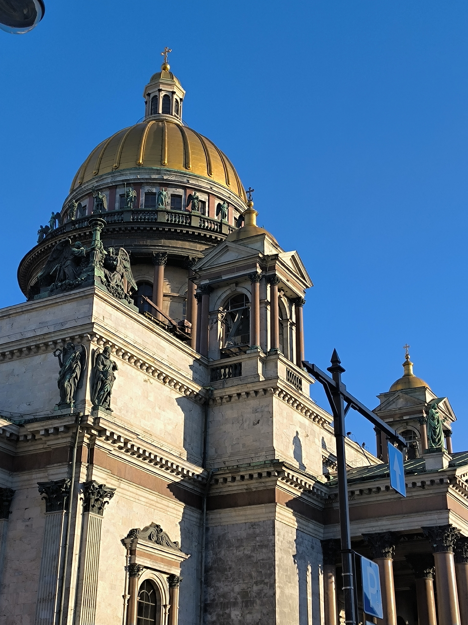 The image shows a low-angle view of Saint Isaac's Cathedral in Saint Petersburg, Russia. The grand building features a large, golden dome topped with a cross, and is adorned with statues and intricate architectural details. The bright blue sky provides a clear backdrop, with the warm sunlight casting shadows on the facade of the cathedral. In the foreground, a street light with a blue directional sign is visible, partially obscuring the view of the building. The overall scene conveys a sense of grandeur and historical significance.