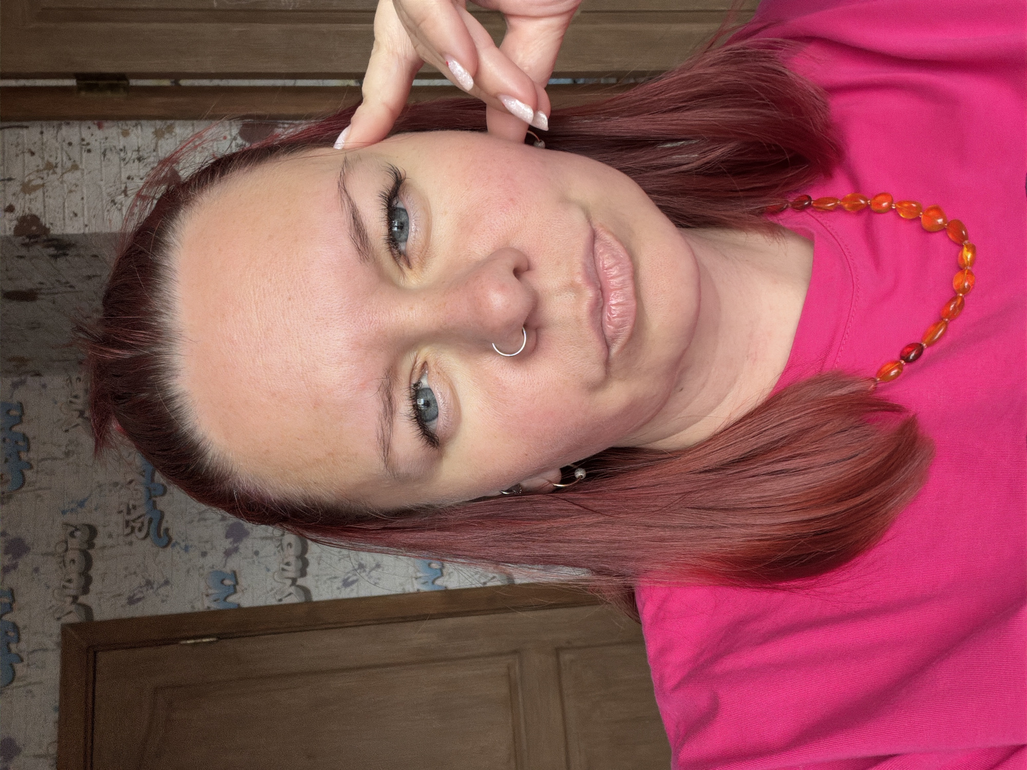 A young, fair-skinned woman with red-tinted hair is visible in a selfie-style photograph. She is wearing a bright pink t-shirt and a necklace of orange-colored beads. A silver nose ring is in her left nostril. She's touching her forehead with her right hand, and her fingernails are decorated. The background includes a wooden door and patterned wallpaper. There is no clear evidence to ascertain that this image was taken in the city of Chaikovsky, Russia. The image depicts a close-up portrait of a person in a domestic setting.