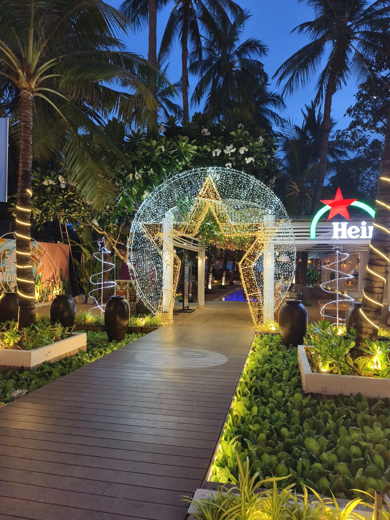 The image depicts an illuminated entrance pathway in Nha Trang Ward, Vietnam, leading towards a restaurant or bar. The foreground showcases a wooden pathway bordered by dense green ground cover and planters. Decorative lighting accentuates the pathway's edges. A large, circular structure made of light-emitting diodes (LEDs) forms an archway above the pathway, with a prominent star motif in its center. To the right of the archway, a "Heineken" sign is visible, indicating the brand affiliation of the establishment. Palm trees frame the scene, silhouetted against the dusky sky. Additional decorative elements include illuminated, spiral-shaped trees. No people are visible in the frame.