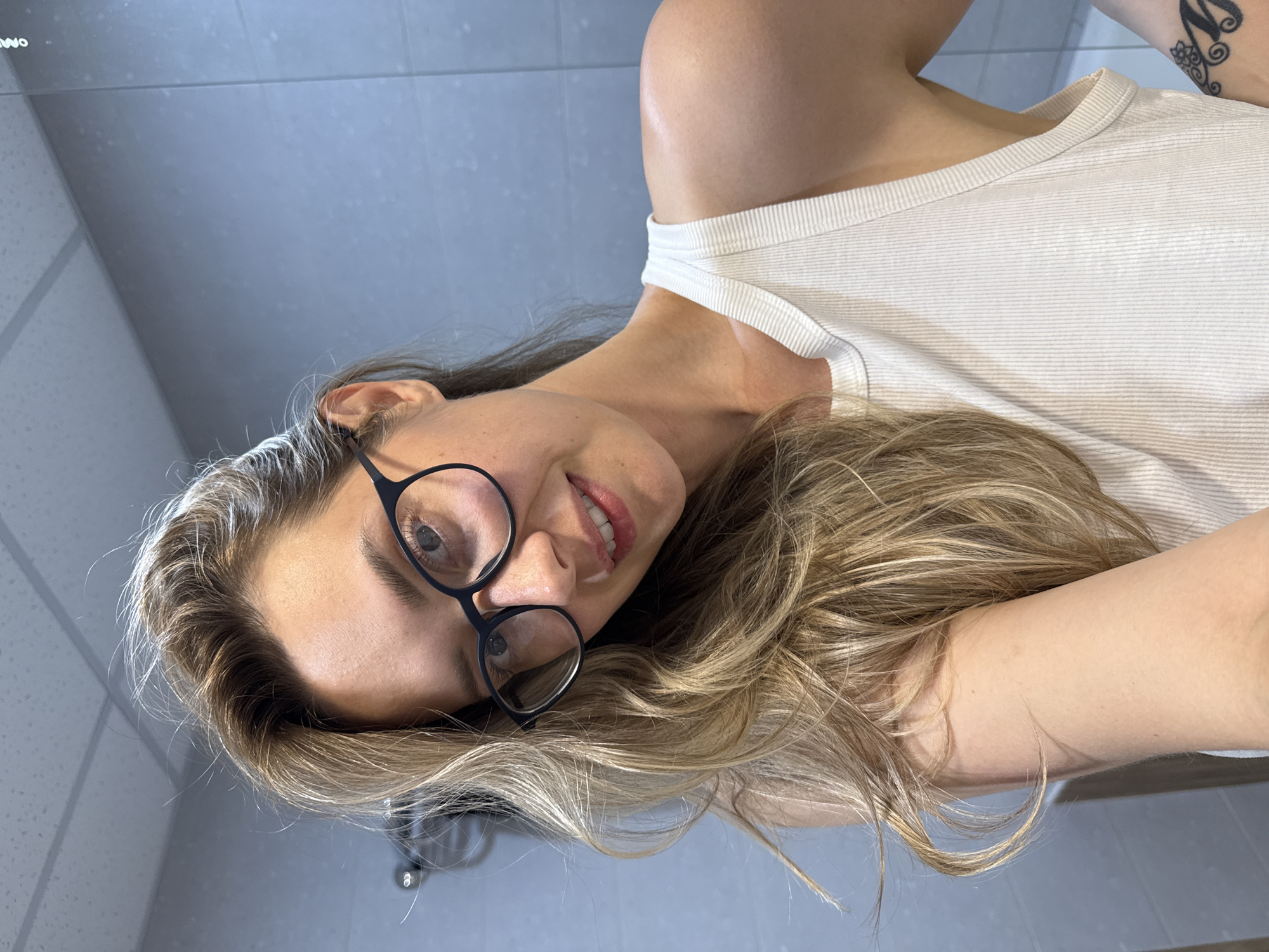 A young woman with blonde hair and glasses is taking a selfie in a bathroom. She is wearing a white tank top. The background is a tiled wall and a ceiling. There is a reflection in the mirror. The image is taken in the city of Nebug, Russia.