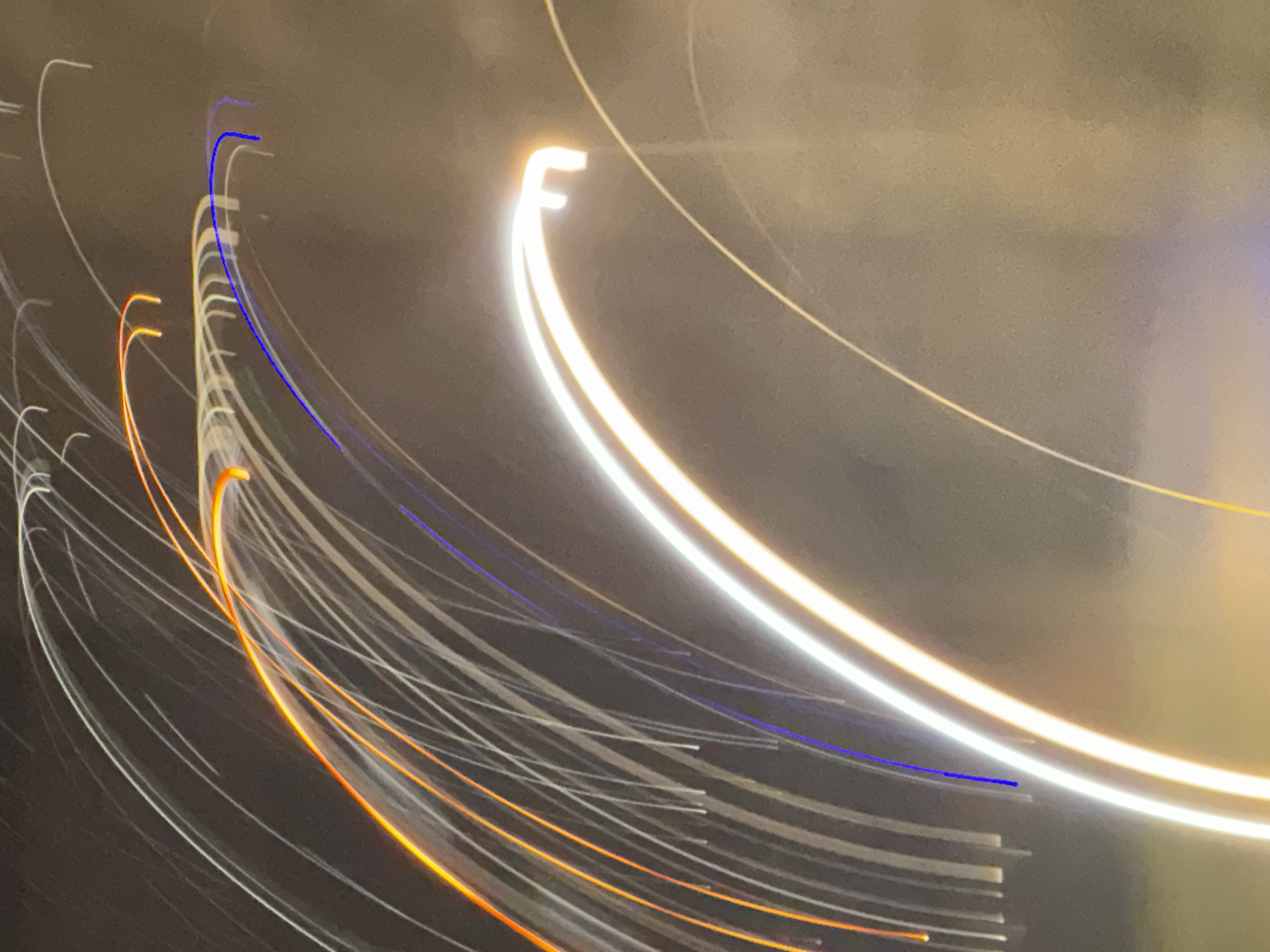 This image captures abstract light trails, likely from long-exposure photography, in the city of Tbilisi, Georgia. The light streaks are primarily in shades of white, orange, and blue, forming graceful arcs and lines against a muted, dark background. The background appears to be a smoky or foggy atmosphere, possibly at night or during twilight. There are no discernible people, subjects, or specific objects in the image, only the dynamic interplay of light that suggests movement and energy. The scene's abstract nature makes it difficult to pinpoint a precise location, but the lighting conditions and the presence of light trails are characteristic of urban environments at night.