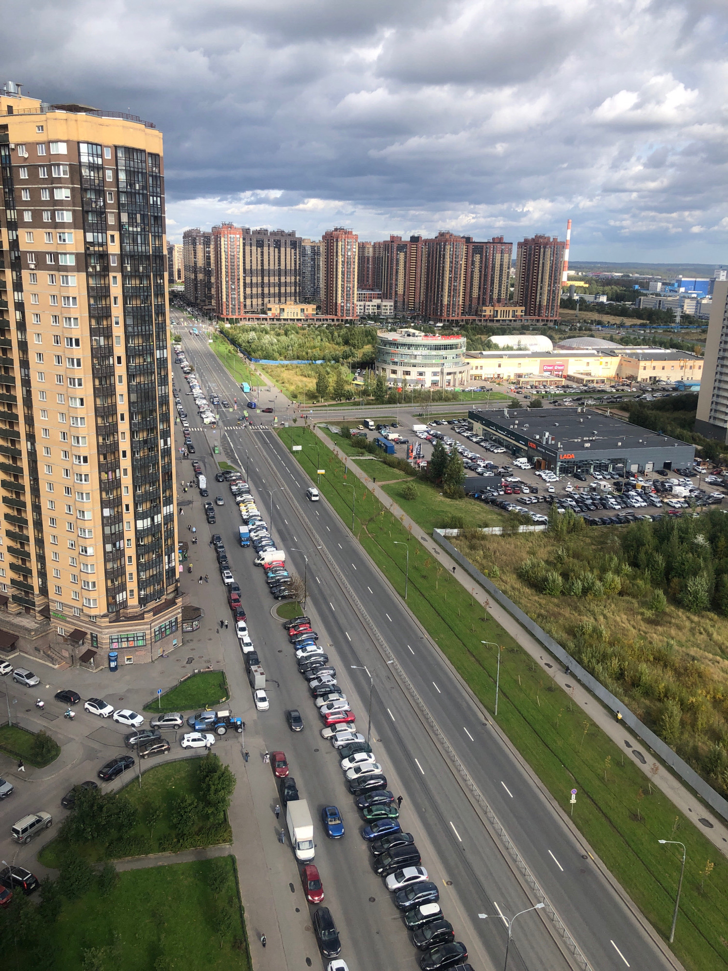 This is an aerial, high-angle, daylight, exterior shot of a suburban environment in Vsevolozhsk, Russia. The scene is dominated by a multi-lane road running diagonally across the frame from the bottom left to the top right. Numerous parked cars line both sides of the road, with a particularly dense concentration on the right side.
To the left of the road, a very tall, multi-story residential building with a light yellow and brown facade occupies the foreground and mid-ground. Its numerous windows are visible. Adjacent to this building, there is a sidewalk and a small green space.
Further down the road on the right, a large, dark-roofed building with a visible "LADA" sign indicates a car dealership. A large parking lot in front of this dealership is filled with cars. Beyond the dealership, a wide expanse of green grass and trees extends into the background, with more distant buildings and a red and white striped chimney visible on the horizon. The sky is cloudy with patches of blue.
Several individuals are visible walking on sidewalks and near parked cars. There are also a few vehicles in motion on the road, including cars and a white van. The overall impression is one of a bustling suburban area with significant car traffic and modern residential and commercial structures.
