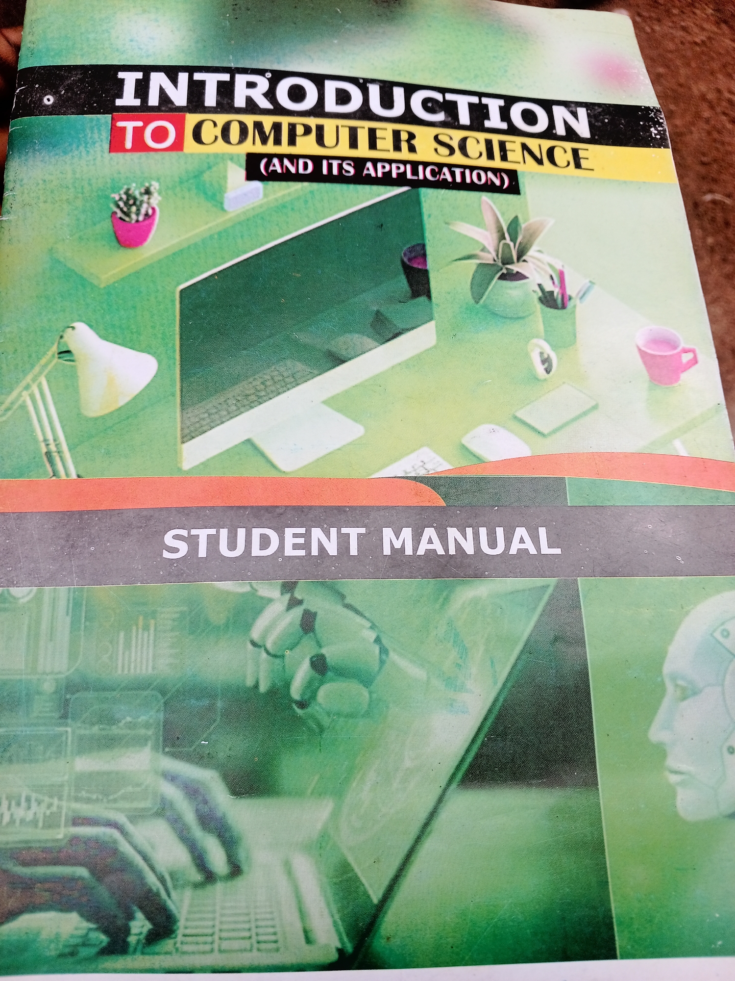 This is a medium shot of a book cover. The cover features a graphic design with a green background that appears to be a stylized representation of a desk with computer equipment. On the desk, there are several objects, including a desk lamp, a computer monitor with a keyboard, a pink plant pot with a small succulent, another plant in a green pot, a pink mug, and what looks like a tablet and a computer mouse. The design also incorporates images of robotic hands typing on a keyboard and a profile of a robot's head.
The text on the cover is prominently displayed. At the top, in large white letters on a black background, is the word "INTRODUCTION". Below this, in red letters on a yellow background, are the words "TO COMPUTER SCIENCE". Further down, in white letters on a black background, is the text "(AND ITS APPLICATION)". At the bottom, in large white letters on a dark gray background, is the text "STUDENT MANUAL".
The overall impression is one of a textbook or study guide related to computer science, with a modern and technological theme. There are no people visible in the image, only graphical representations of hands and a robot head. The lighting appears to be artificial, suggesting an indoor setting. The image itself is somewhat grainy and shows signs of wear, with some dust specks visible on the surface. No specific time of day, weather, or emotional cues are discernible. The provided location context of Jalingo, Nigeria, does not provide any direct visual cues within the image itself.