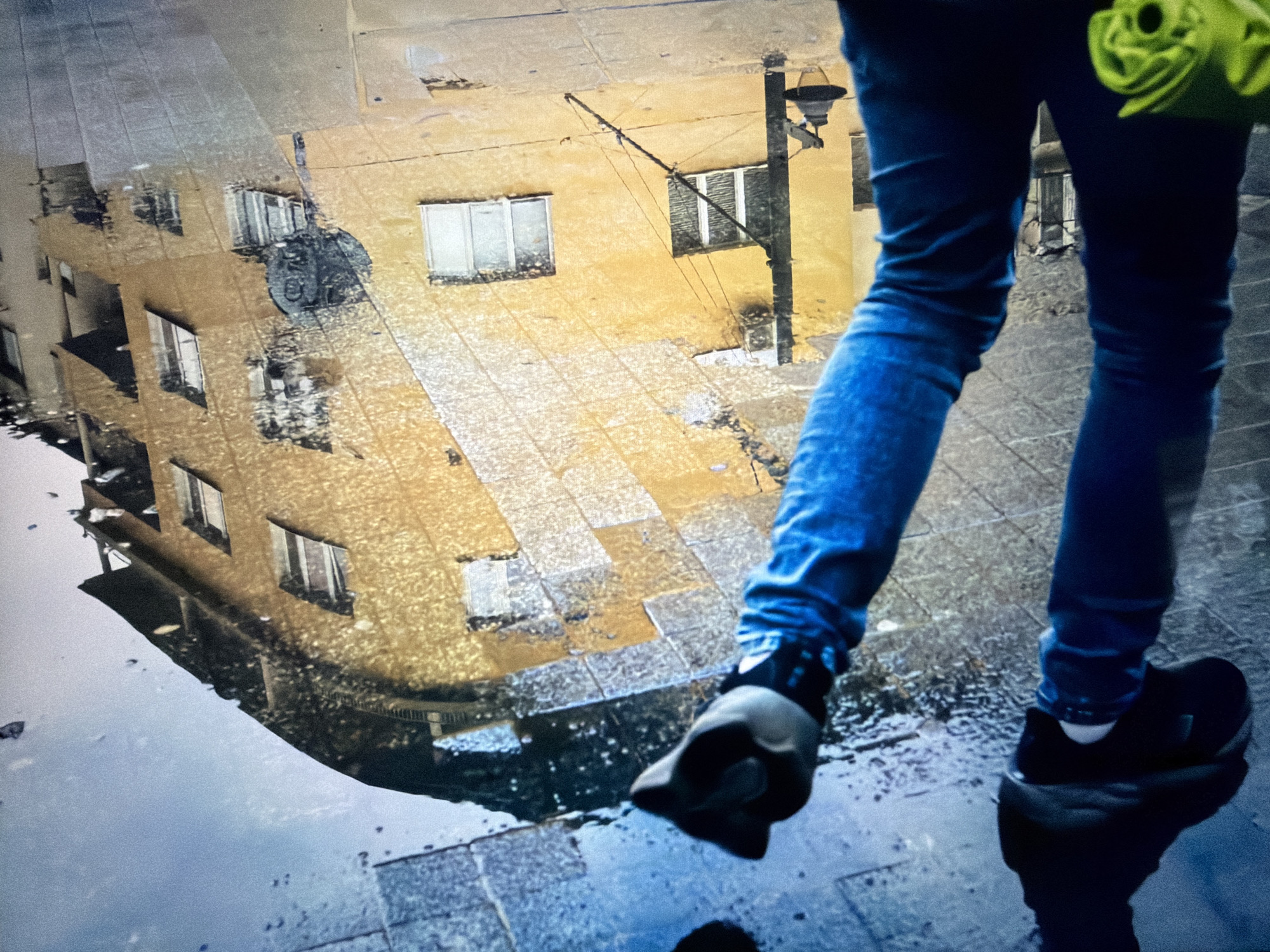 The scene depicts a pedestrian walking on a wet, tiled pavement in the city of Burgas, Bulgaria, with a large puddle reflecting the surrounding urban environment. The lower torso and legs of a person, clad in fitted blue denim jeans and dark athletic shoes, are visible. The left hand, partially obscured by a dark glove or wristband, extends forward and holds an indistinct bright green object. The person's right foot is forward, with its reflection visible in the water, while the left foot is slightly behind, appearing to step into the puddle.
The puddle occupies the mid-ground and foreground, creating a clear reflection of a multi-story building. This building has a distinctive light yellow-ochre facade, punctuated by numerous rectangular windows with white frames. A prominent black streetlamp post, featuring a traditional-style lantern and attached wires, stands against the building wall within the reflection. Near one of the reflected windows, a dark, circular object, potentially a bicycle wheel, is affixed to the building's exterior. The reflected ground surface shows a pattern of square or rectangular pavement tiles, appearing wet and textured. The overall impression is an urban setting after rainfall, characterized by reflections and muted lighting.